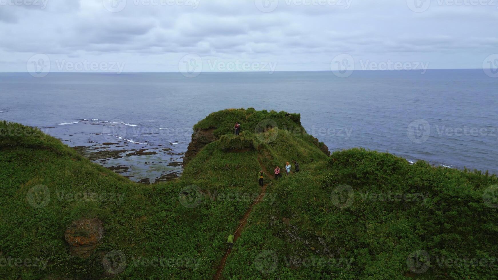 Top view of tourists on rocky coast trail with green grass. Clip. Beautiful landscape of rocky coast with green grass and tourists on hike. Tourists ride on edge of rocky coast overlooking sea on photo