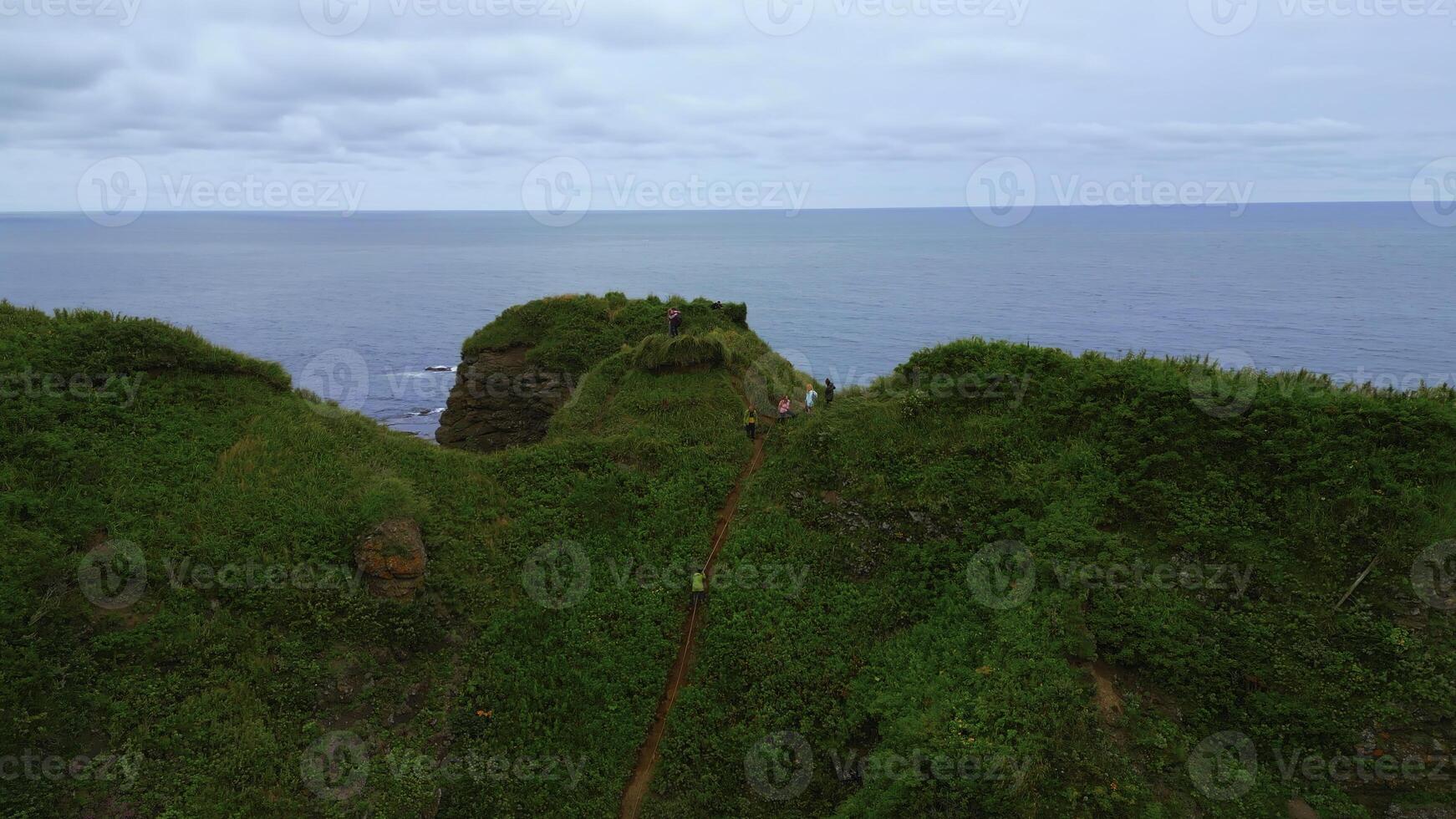 Top view of group of people standing on edge of coast. Clip. Tourists are standing on rocky shore overlooking sea horizon. Tourists on rock with beautiful grass by sea on cloudy day photo