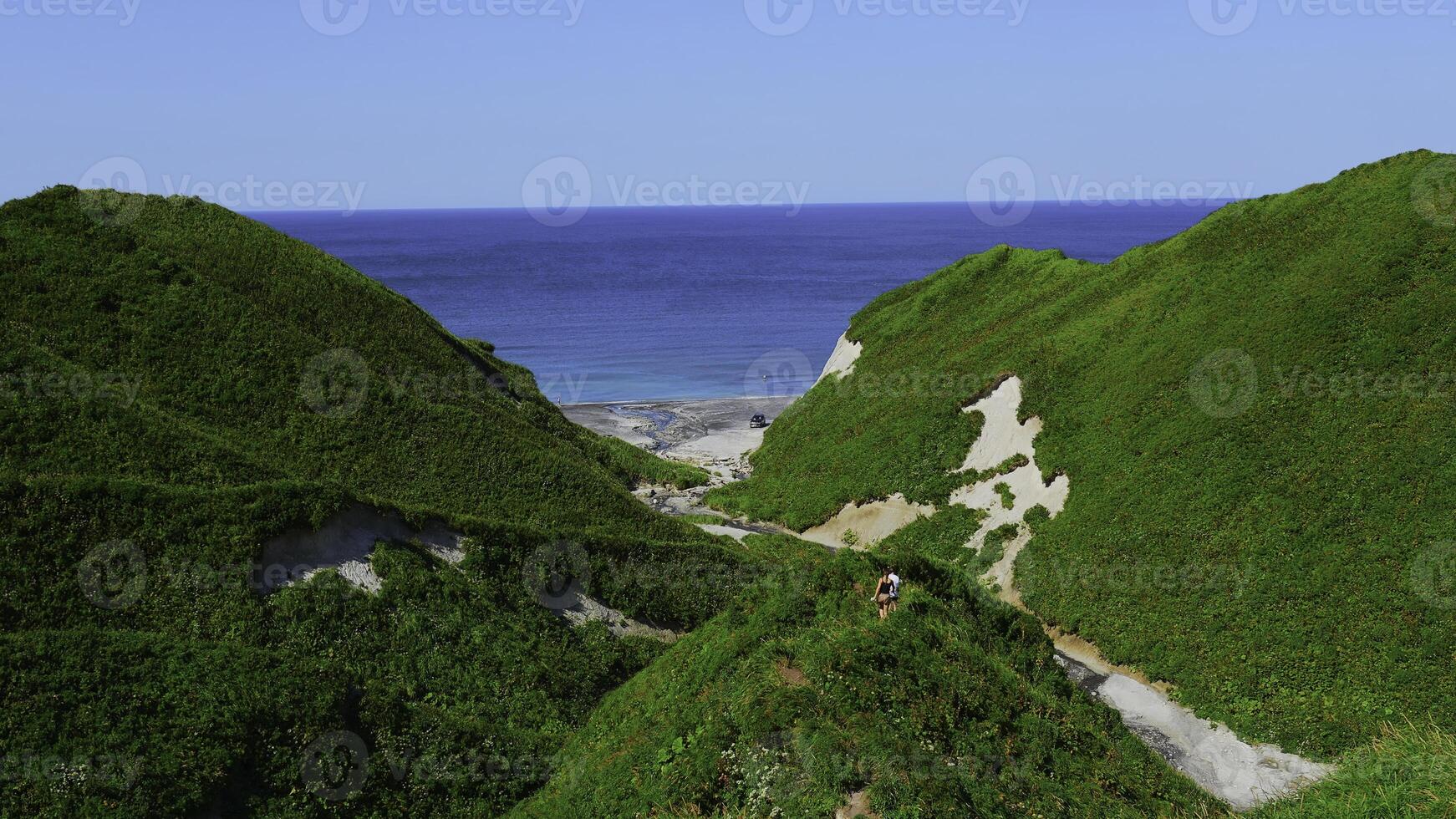 Couple walking along path on hills on background of sea. Clip. Top view of hills with trails and green grass on background of sea coast. Beautiful summer hills by sea on sunny day photo