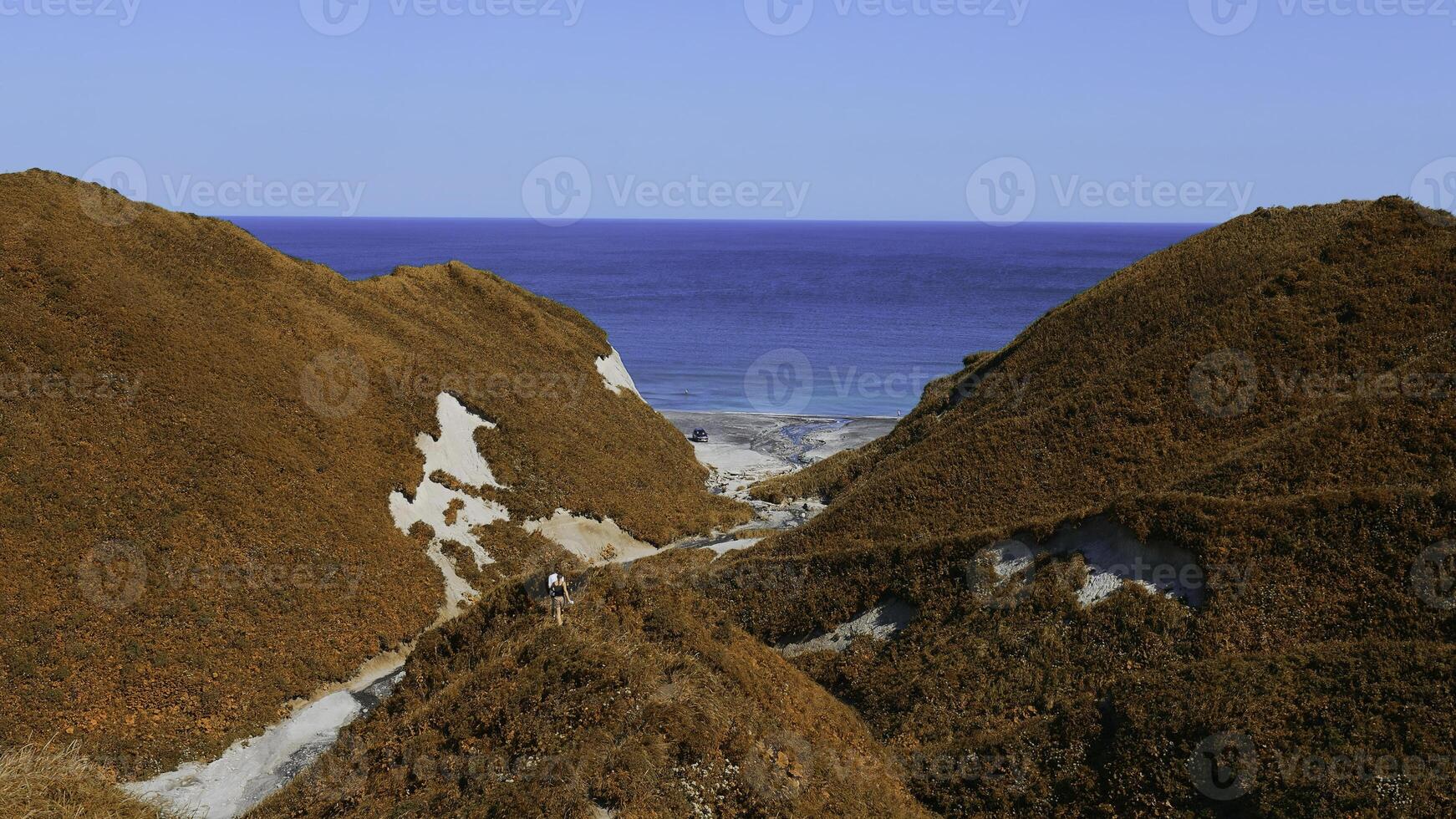 Couple walking along path on hills on background of sea. Clip. Top view of hills with trails and green grass on background of sea coast. Beautiful summer hills by sea on sunny day photo