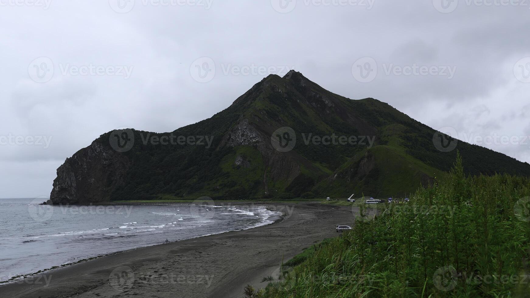 View of beautiful beach with green grass and mountain. Clip. Picturesque landscape with green grass on background of sea coast with mountain. Coast of sea with sand and rocky mountain on cloudy day photo
