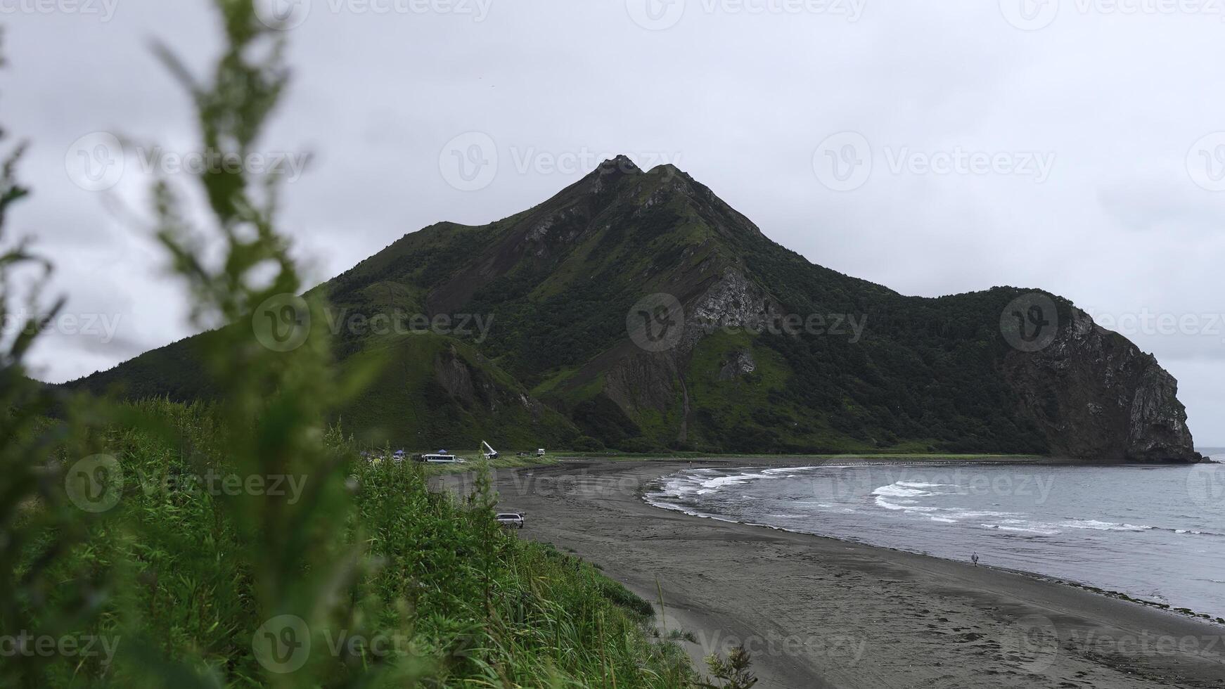 View of beautiful beach with green grass and mountain. Clip. Picturesque landscape with green grass on background of sea coast with mountain. Coast of sea with sand and rocky mountain on cloudy day photo