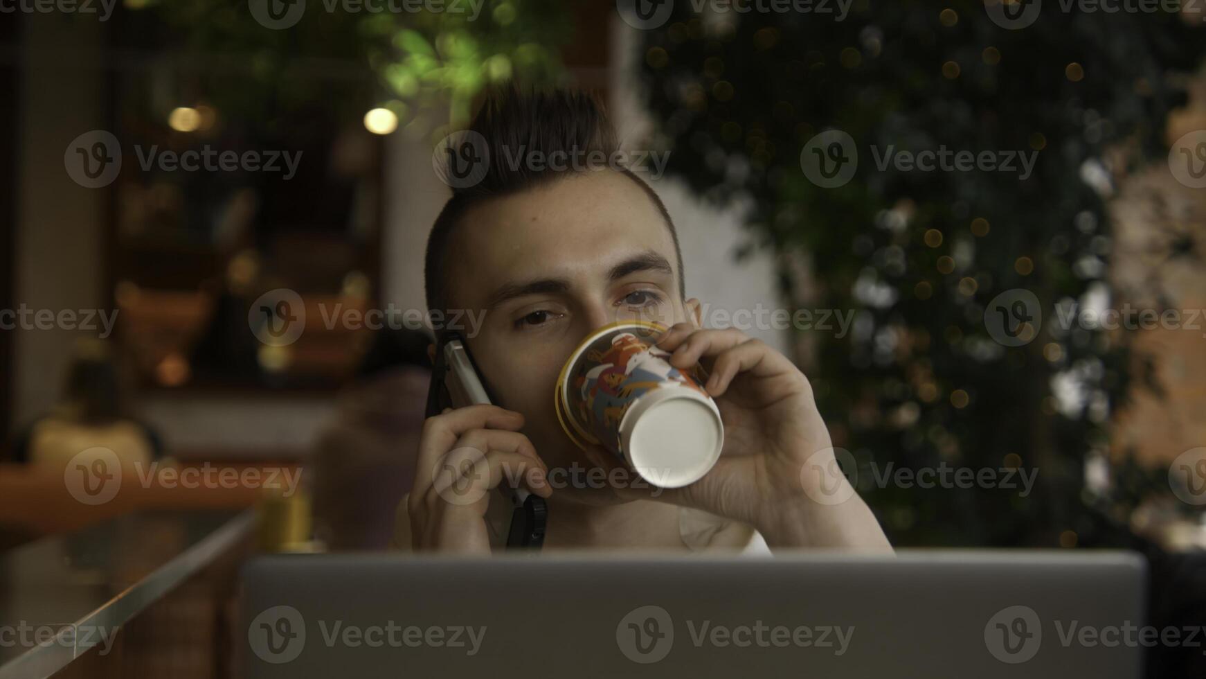 Young man is working on laptop in cafe and talking on phone. Stock footage. Young freelancer is working on laptop and answering calls. Remote work with laptop and cafe calls photo