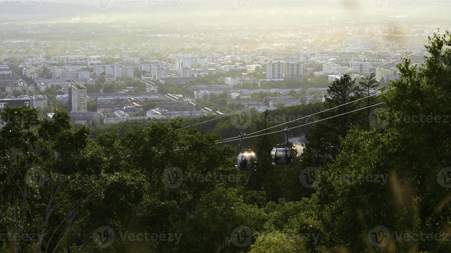 Beautiful view of cable car with view of town in summer. Clip. Green forest on slope with moving cable ways on edge of town. Beautiful city in valley with cable car on sunny summer day photo