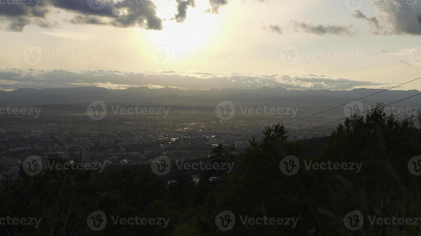 Beautiful view of city with cable car. Clip. Landscape of city in valley on background of mountain horizon with sun. Cloudy weather with sun is over valley with town and cable car photo