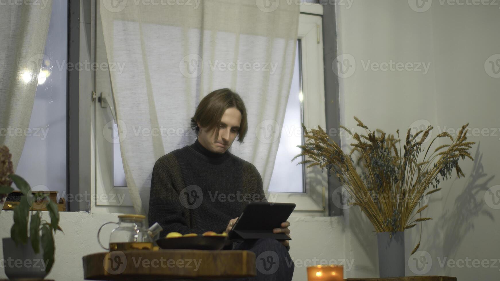Young man drinks tea and works on tablet in cafe. Media. Student is relaxing with tablet and tea in cozy cafe. Handsome student studying on break in cafe photo