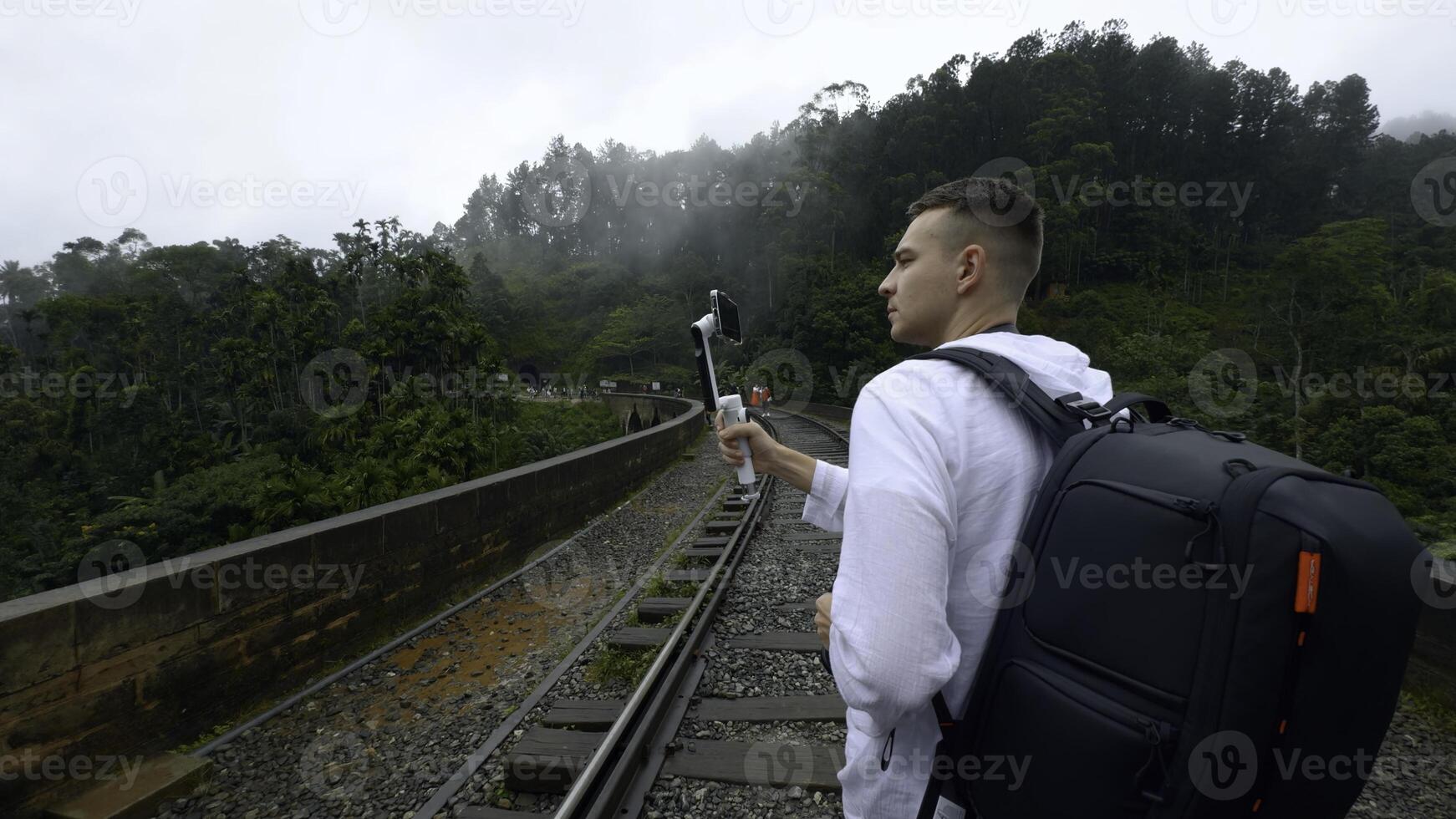 posterior ver de un hombre turista tomando imágenes de montaña boscoso paisaje en teléfono inteligente acción. caminante caminando en vias ferreas. foto