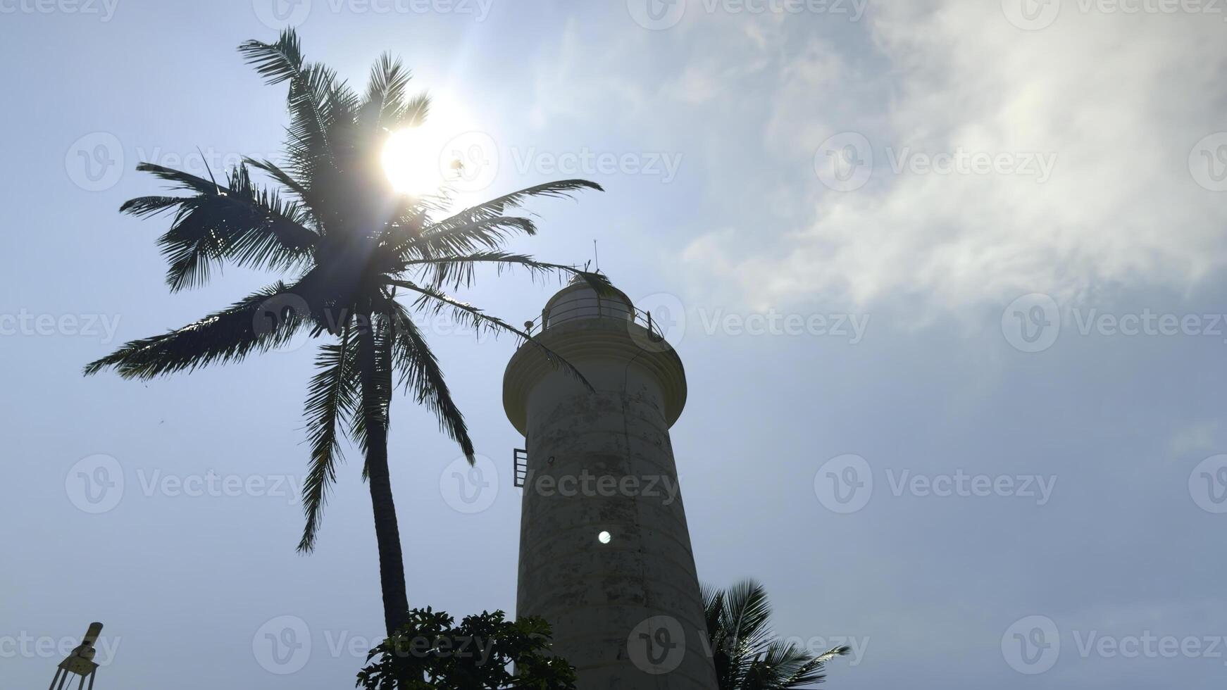Low angle view of a beautiful tower and a palm tree against blue cloudy sky. Action. White lighthouse and palm tree. photo