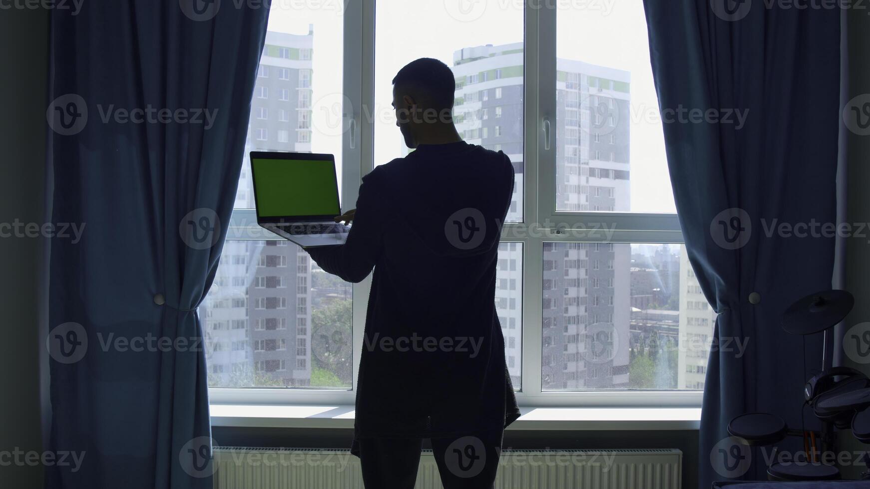 Rear view of a male student studying at home and using his laptop. Media. Man standing by the window and looking at green chroma key screen. photo