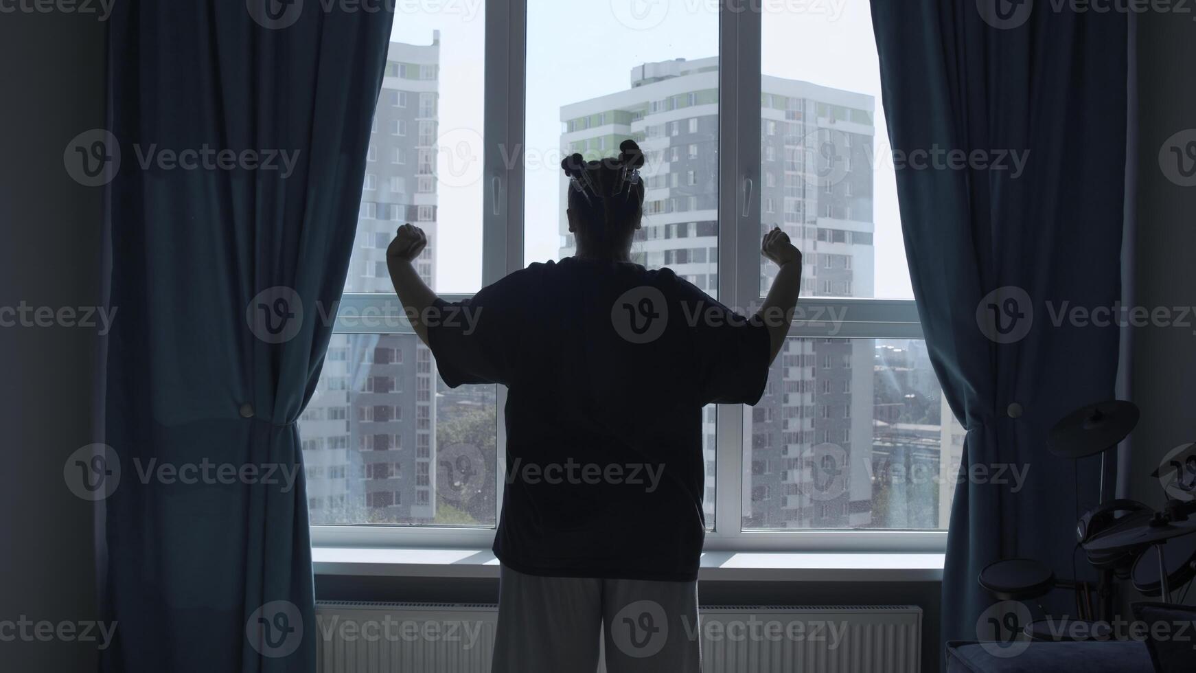 Rear view of a girl in casual clothes in modern bedroom in the morning. Media. Woman looking through the window, raising hands and stretching body muscles. photo