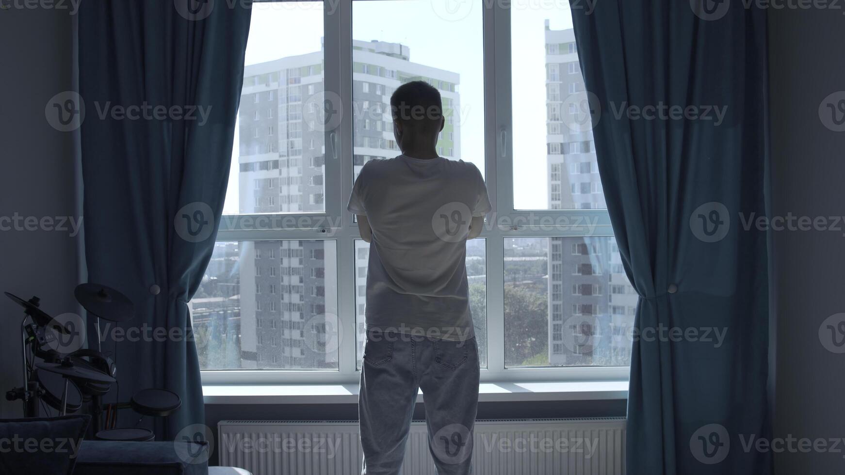 Rear view of a pensive man standing at home by the window and looking at the city. Media. Man in home clothes spending time in reflection in his bedroom. photo