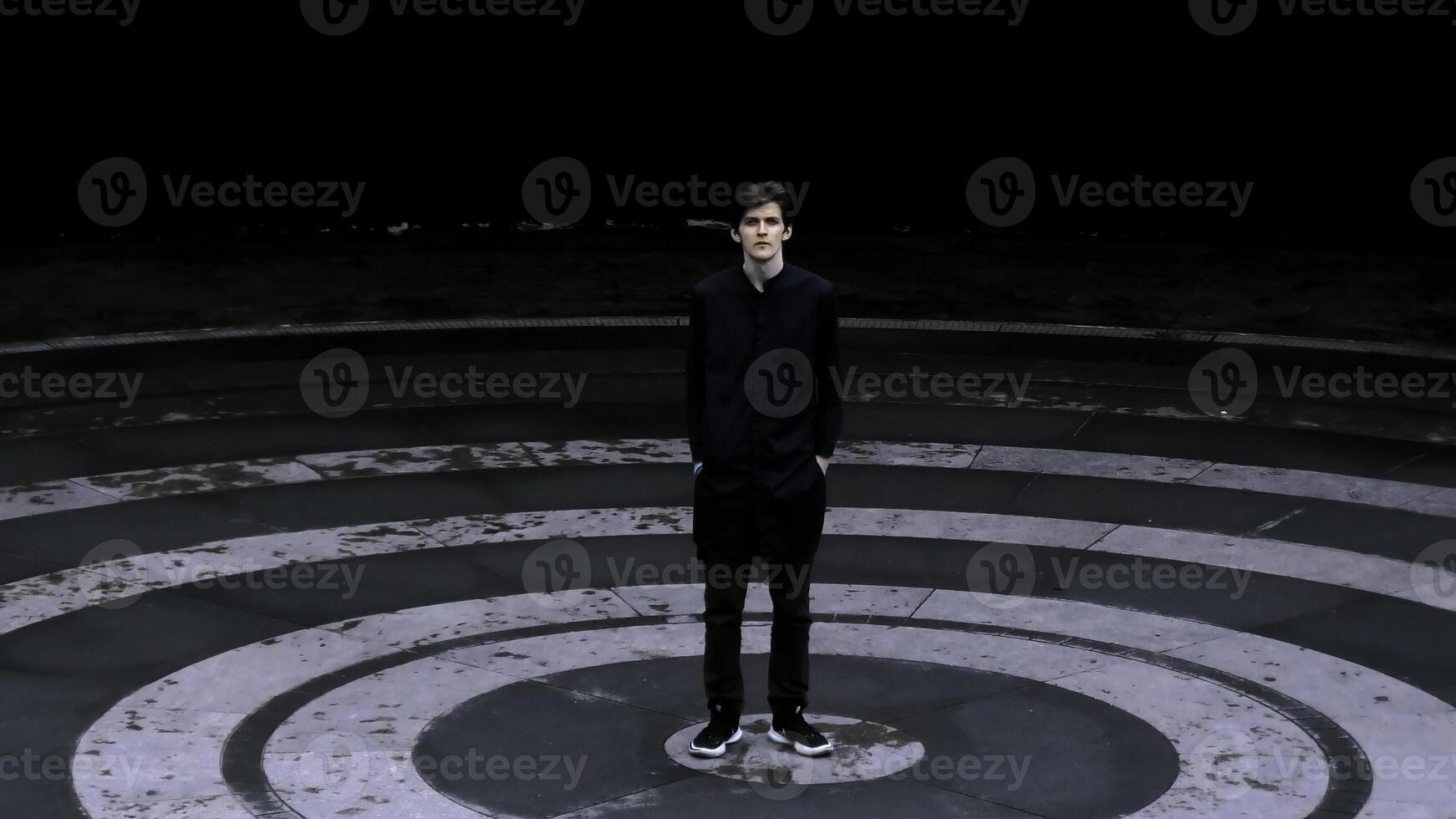 Tall man in black clothes standing in the meddle of amphitheater. Stock clip. Asphalt colored in black circles. photo