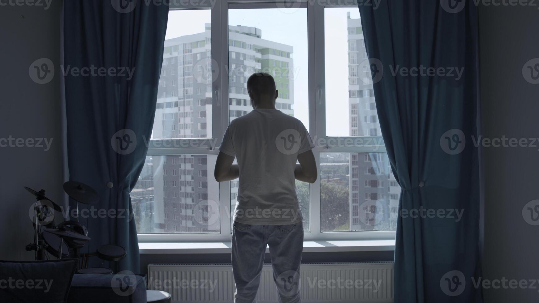 Rear view of a pensive man standing at home by the window and looking at the city. Media. Man in home clothes spending time in reflection in his bedroom. photo