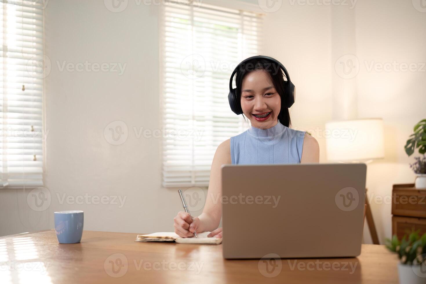 Happy young woman asian in headphones looking at laptop making note, girl student talking by video conference call, online training photo