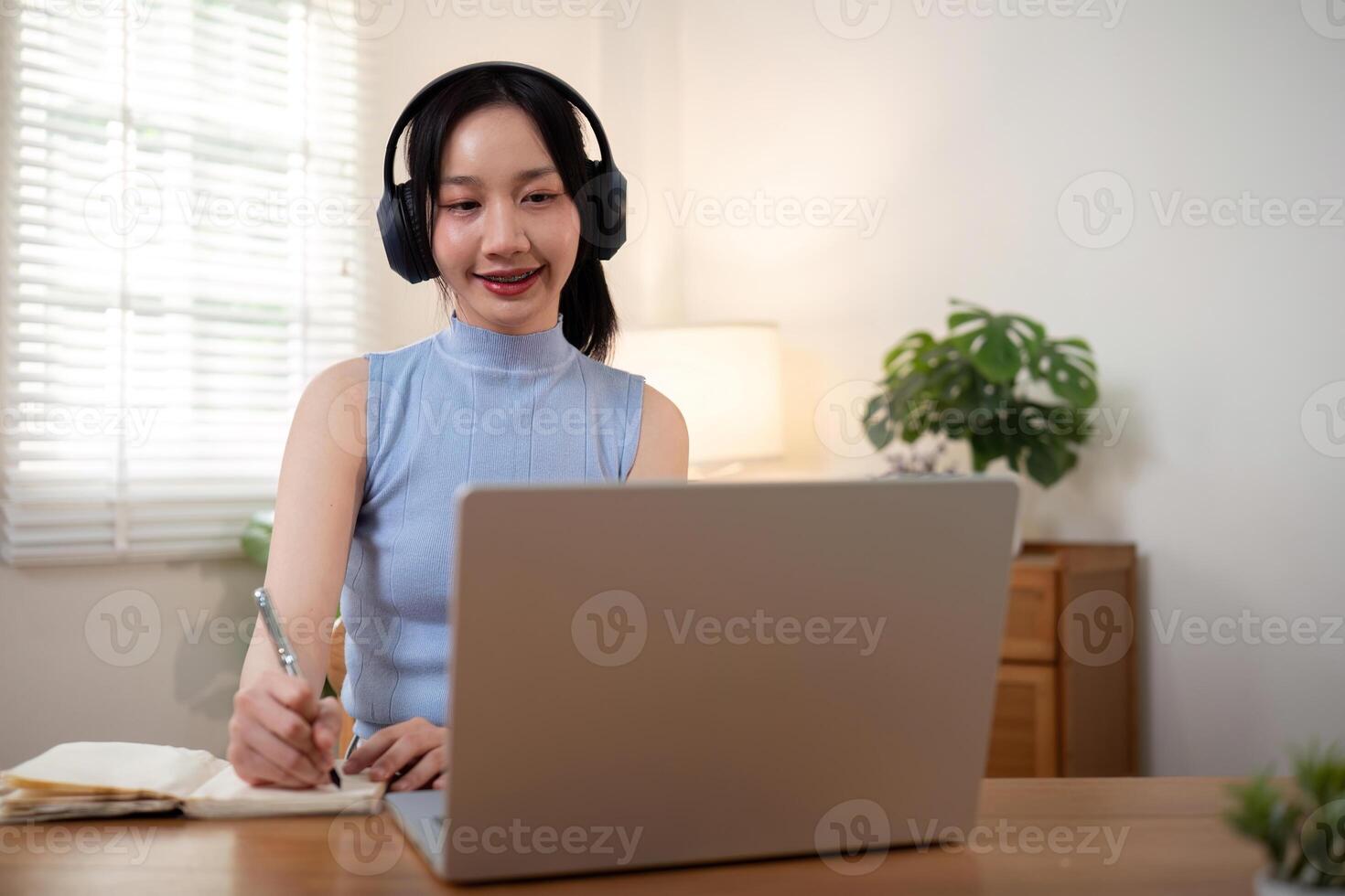 Happy young woman asian in headphones looking at laptop making note, girl student talking by video conference call, online training photo