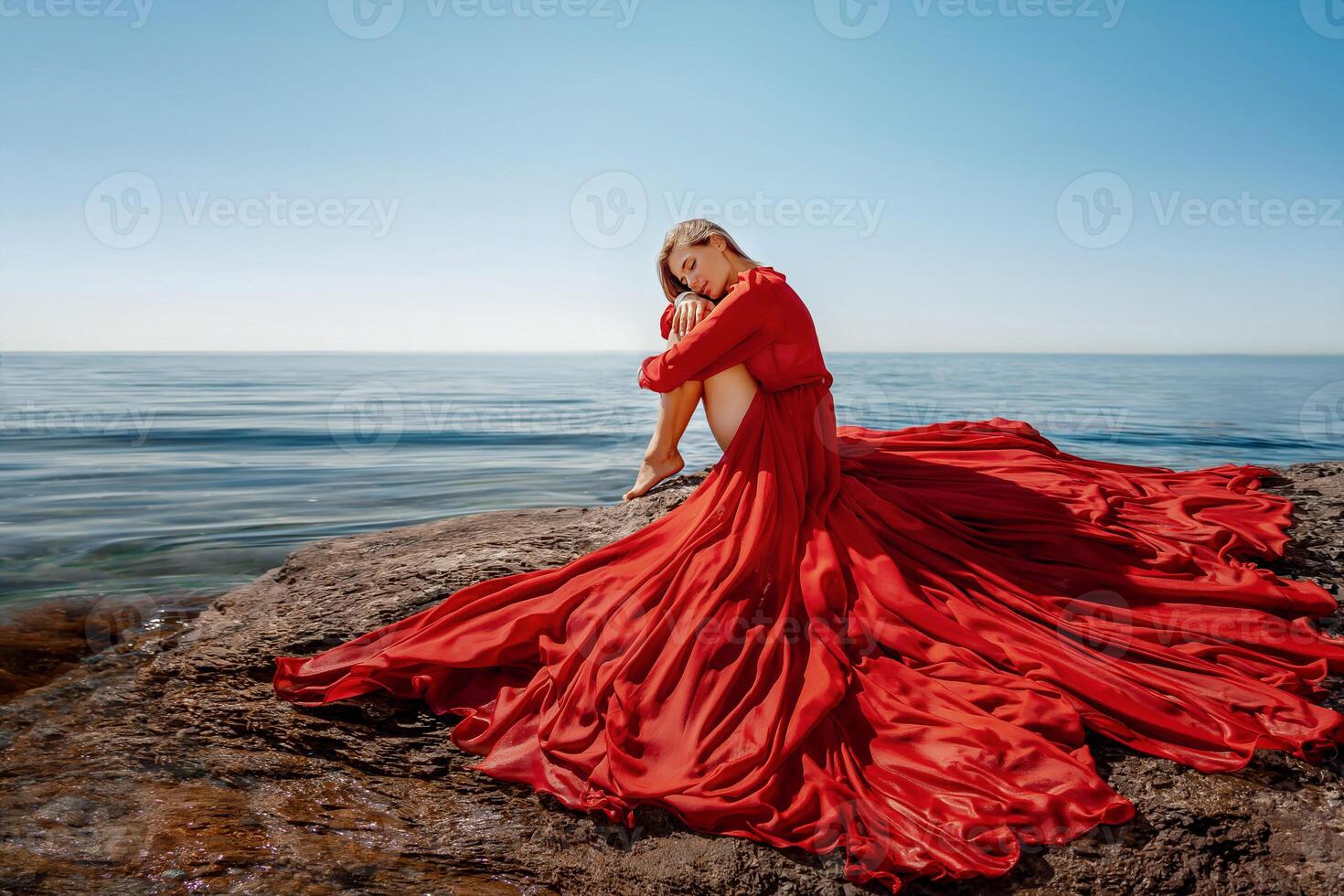 Beautiful sensual woman in a flying red dress and long hair, sitting on a rock above the beautiful sea in a large bay. photo