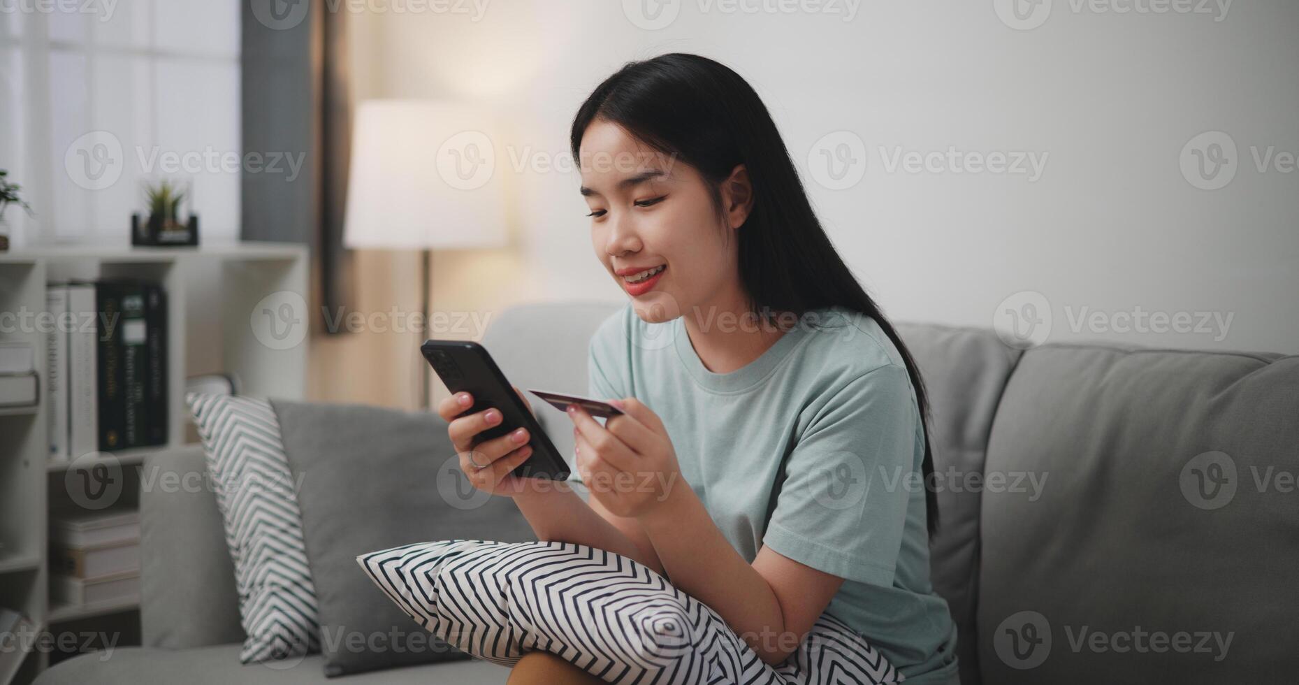 Happy young woman sitting on sofa using credit card with mobile phone for online shopping cashless in living room at home photo