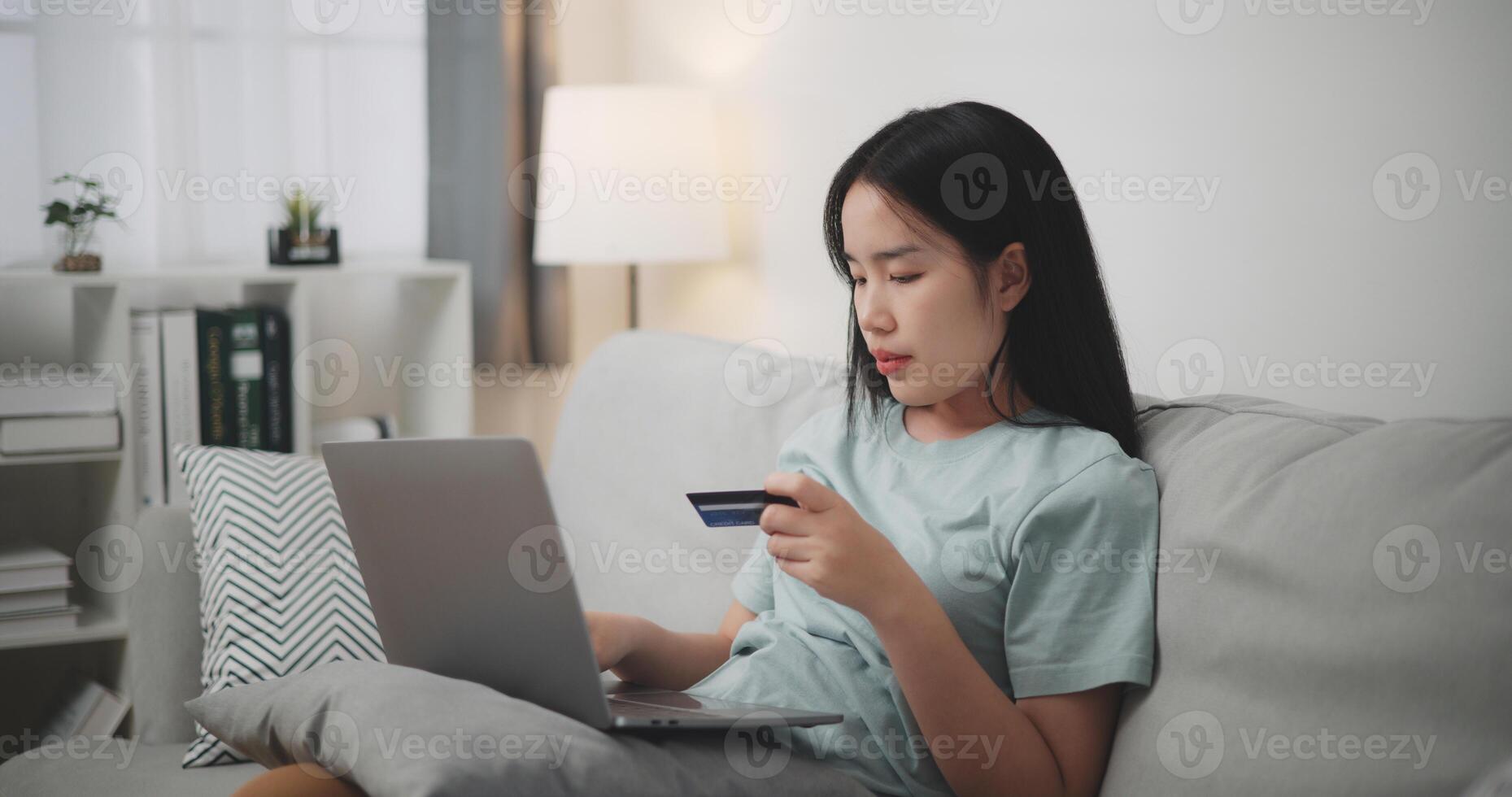 Selective focus, Young asian woman sitting on sofa holding credit card making online payment for purchase in web store using laptop. Ecommerce website payments concept. photo