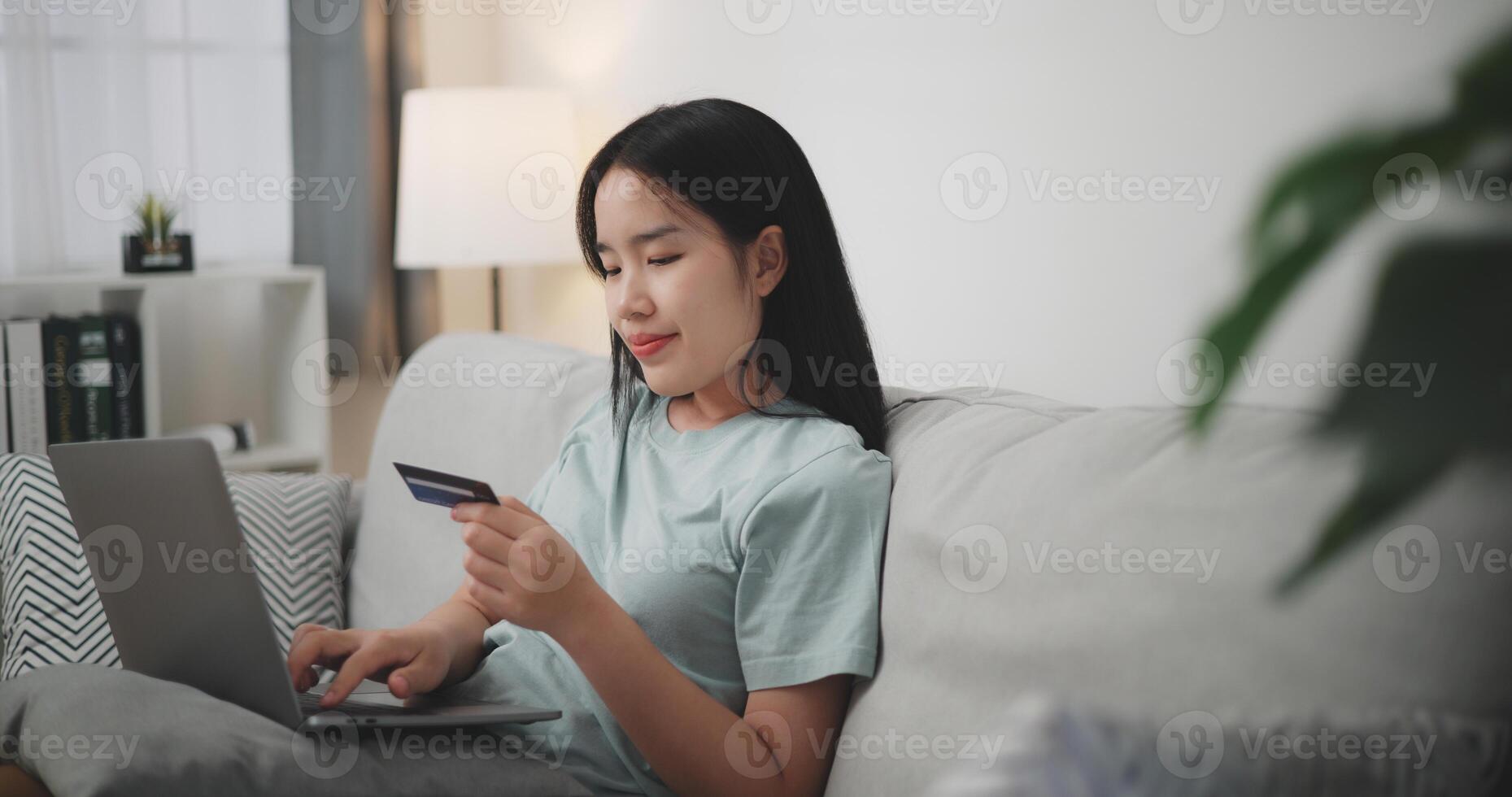Selective focus, Young asian woman sitting on sofa holding credit card making online payment for purchase in web store using laptop. Ecommerce website payments concept. photo