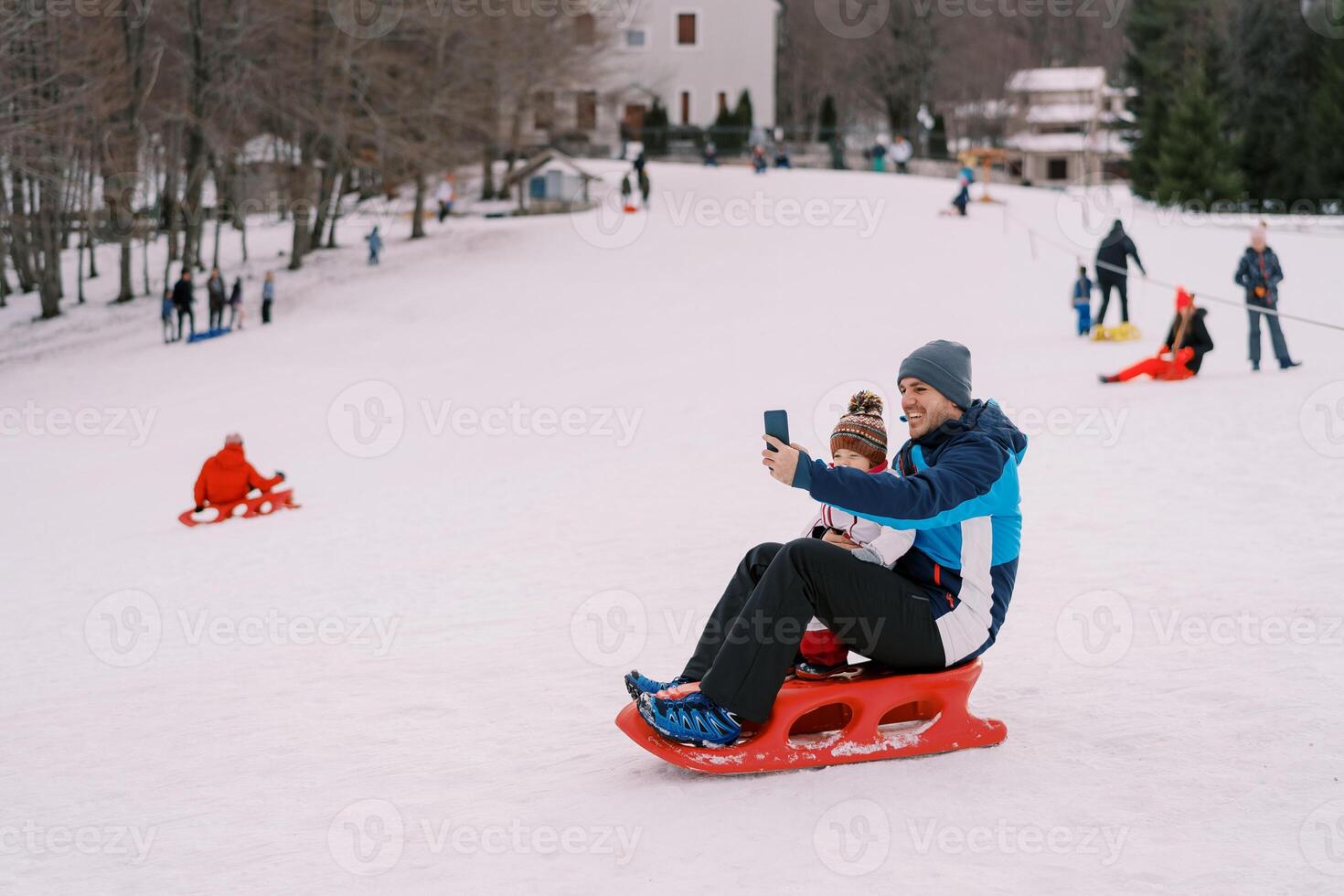 Smiling dad taking smartphone selfie of himself and little kid sitting on sled on snowy hill photo