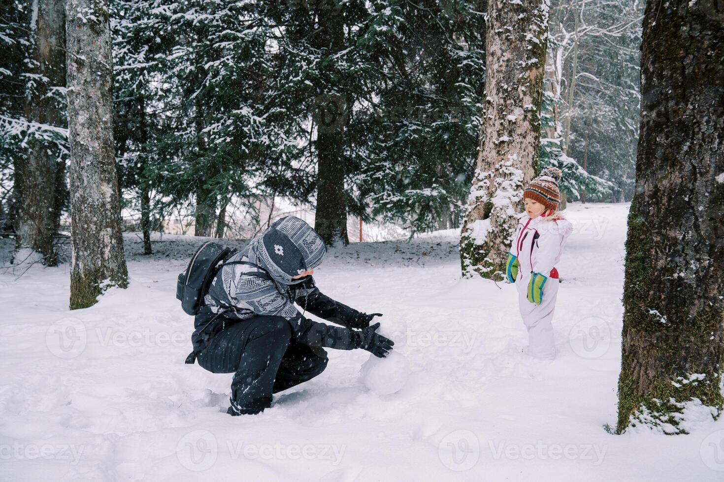 Little girl stands in a snowy forest and looks at her mother making a snowman while squatting photo