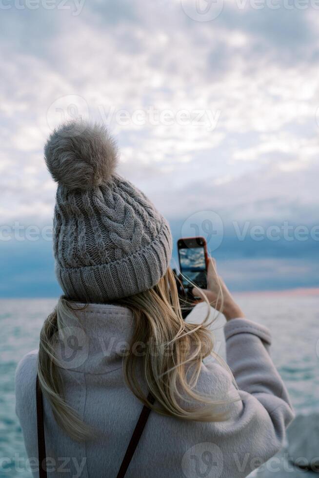 niña soportes en el apuntalar y dispara el puesta de sol terminado el mar con un teléfono inteligente espalda ver foto
