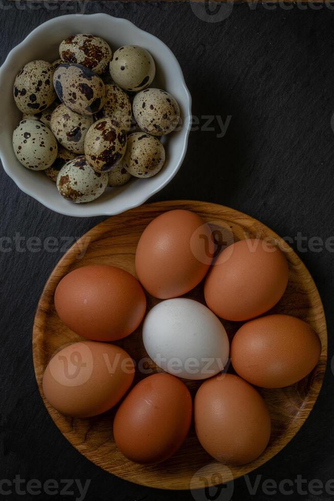 Fresh chicken and quail eggs in white and wooden plate on stone Selective focus. photo