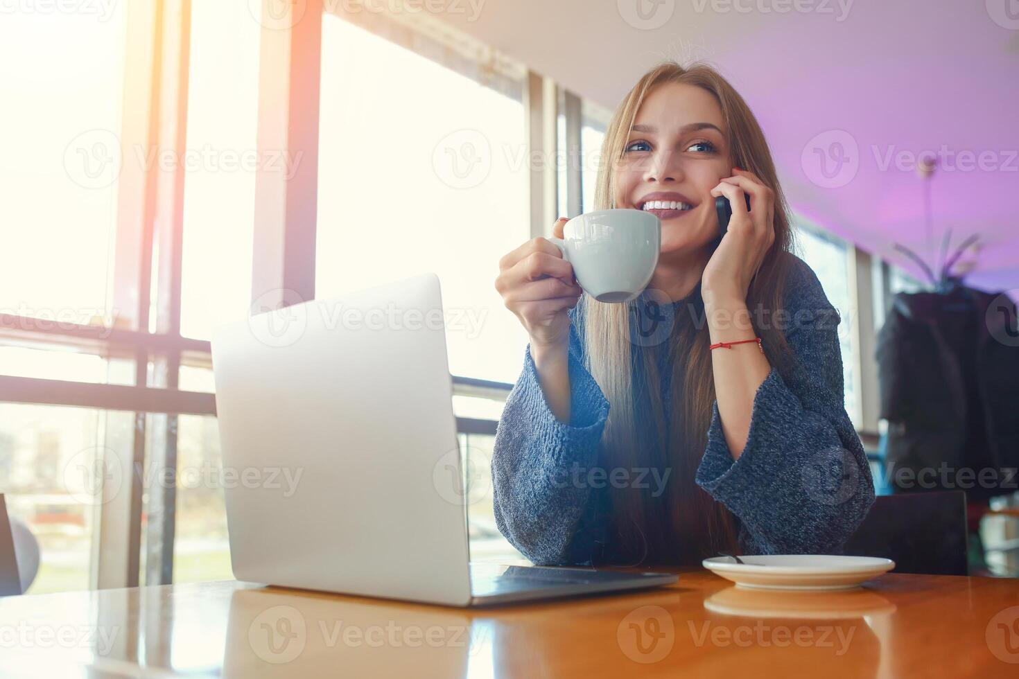 Happy young hipster woman with cup of cofe talking on the phone in at cafe. Laptop on the table. photo