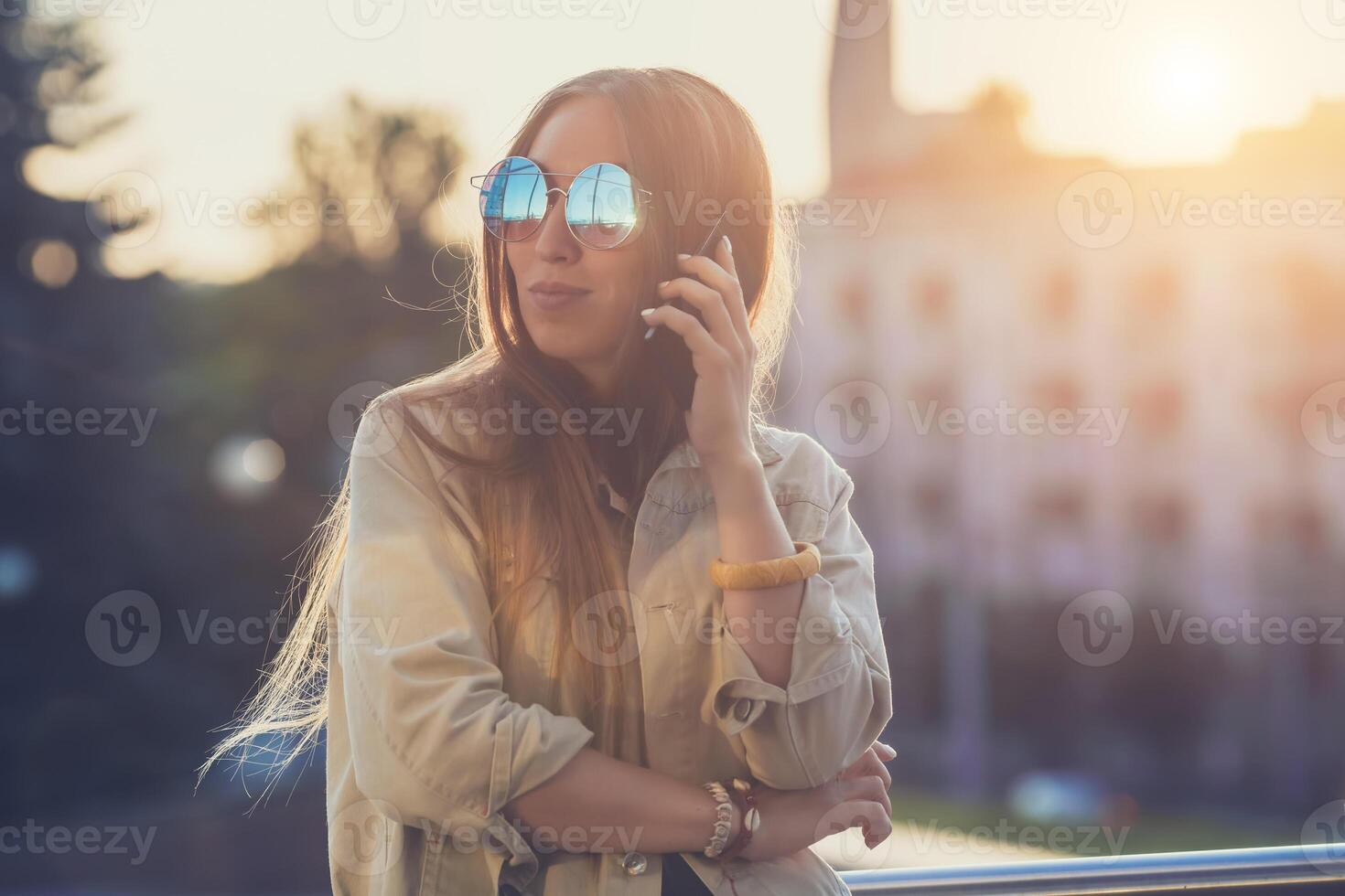 joven elegante bonito mujer, manos participación un teléfono. puesta de sol fondo, soleado día bueno clima, Gafas de sol, frio accesorios. foto