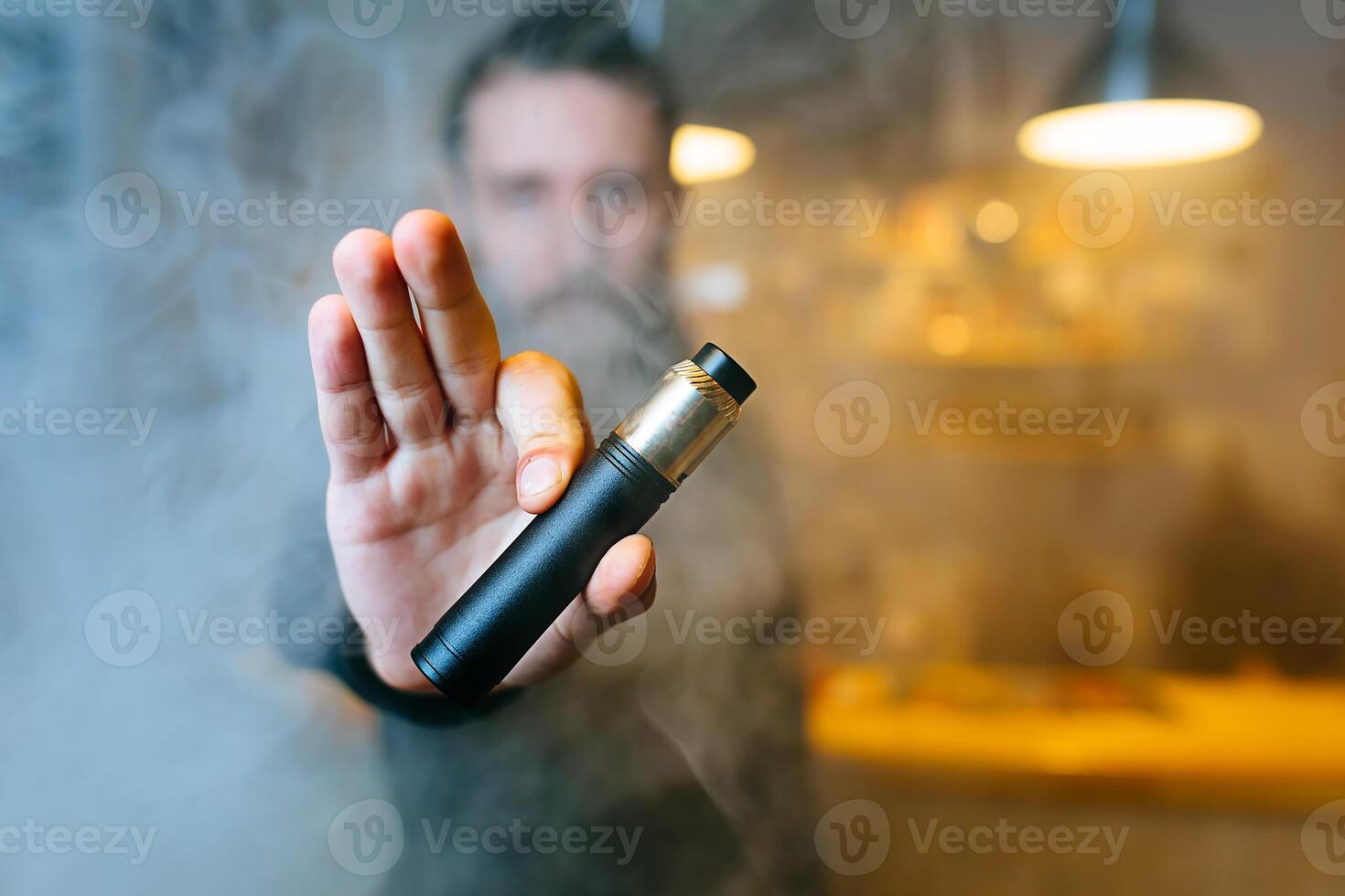 Young beard man show vaping device on his outstretched hand through a cloud of steam. Selective focus. Vaping concept. Copy space. photo