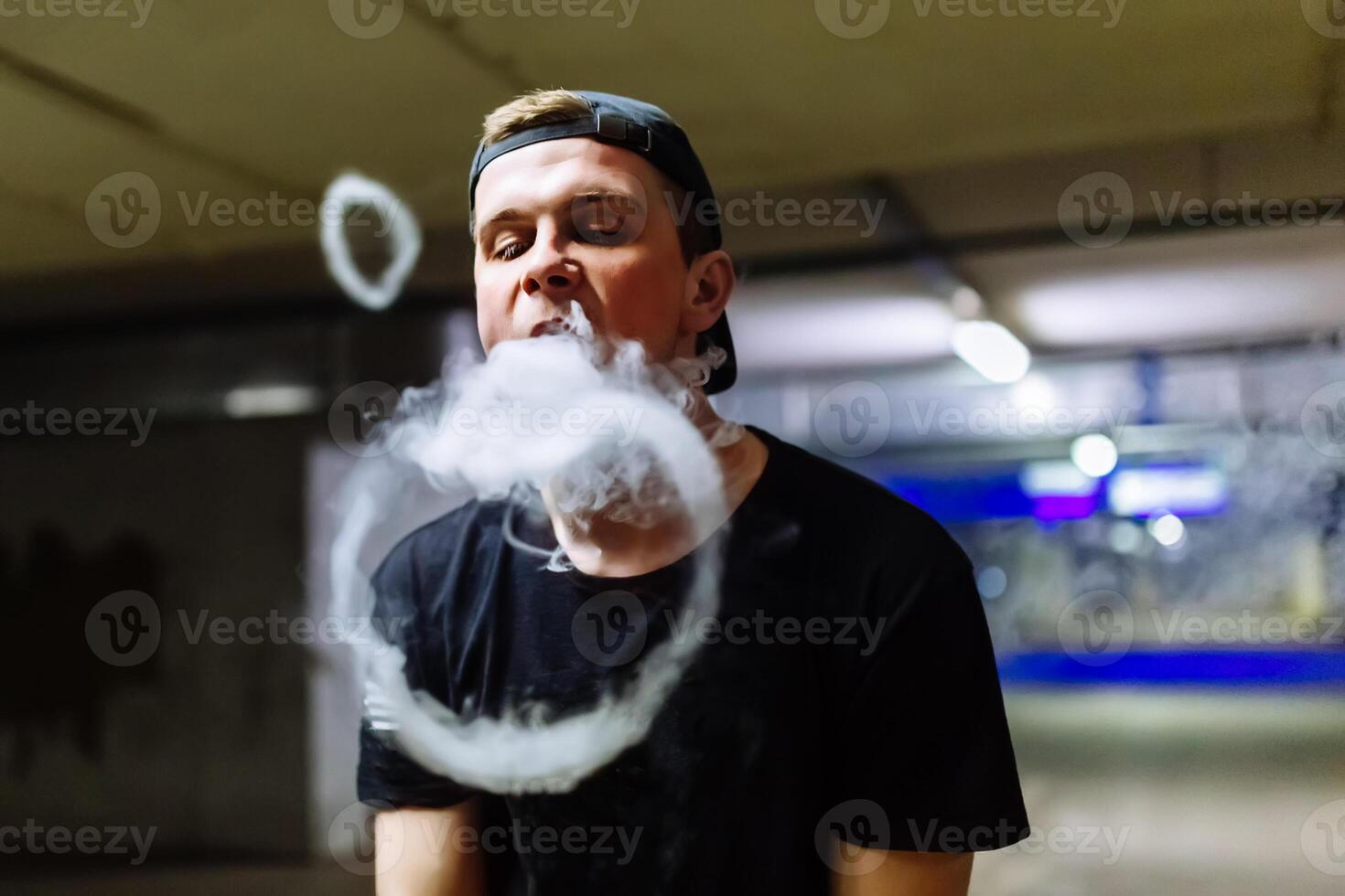 Man in cap smoke an electronic cigarette and releases clouds of vapor performing various kind of vaping tricks photo