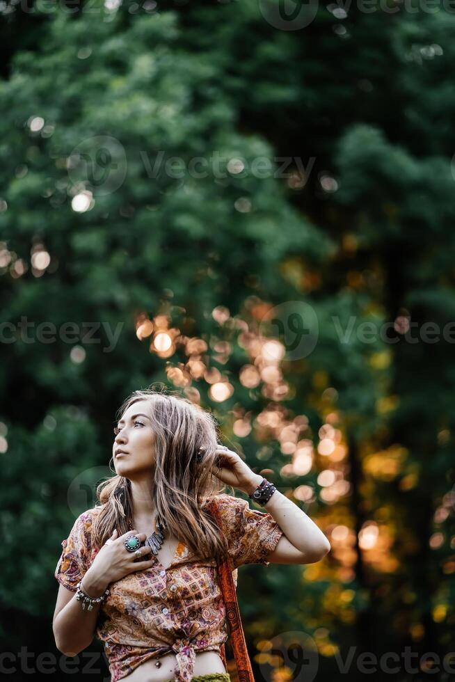 hermosa joven mujer vistiendo estilo boho ropa posando en el rayos de el noche sol, puesta de sol. boho estilo moda, hembra vistiendo plata joyería teniendo divertido en parque al aire libre. foto