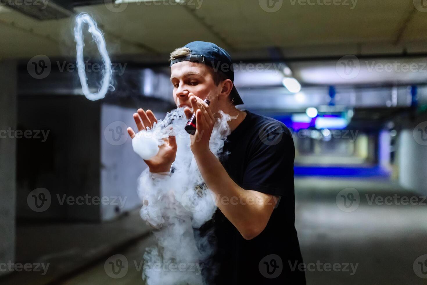 Man in cap smoke an electronic cigarette and releases clouds of vapor performing various kind of vaping tricks photo