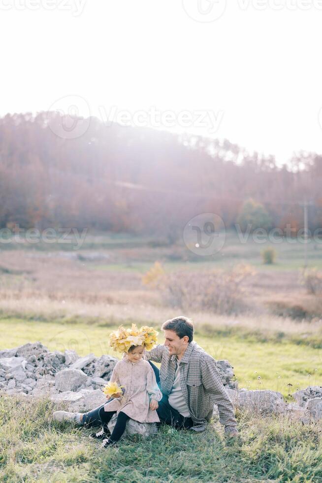 pequeño niña en un guirnalda de amarillo hojas se sienta en un Roca siguiente a su papá en el soleado césped foto