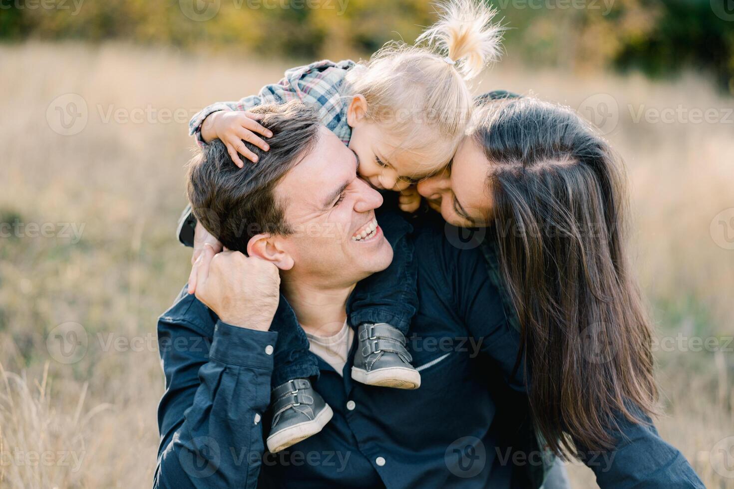 Mom with a little girl on the shoulders of dad kiss him, hugging from behind photo