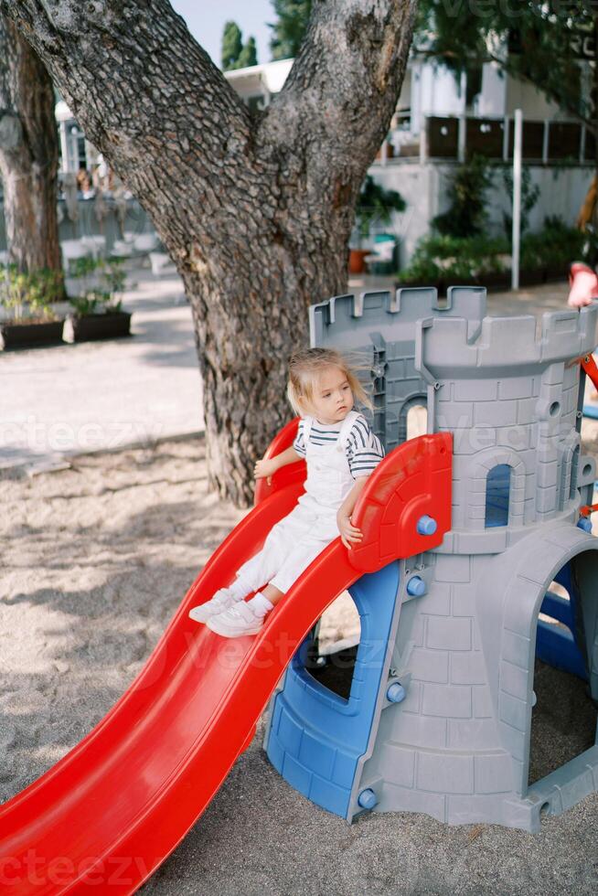 pequeño niña se sienta en parte superior de un deslizar, participación el pasamanos y mirando lejos foto