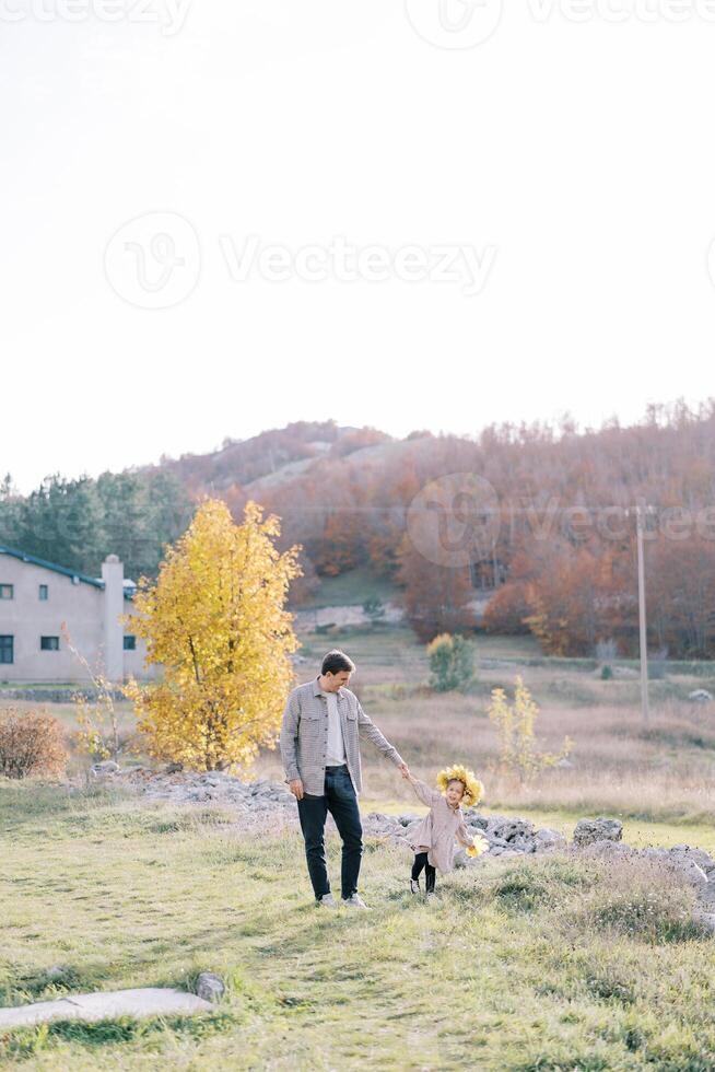 Dad looks at the little girl in a wreath of yellow leaves as he walks holding hands across the sunny lawn photo