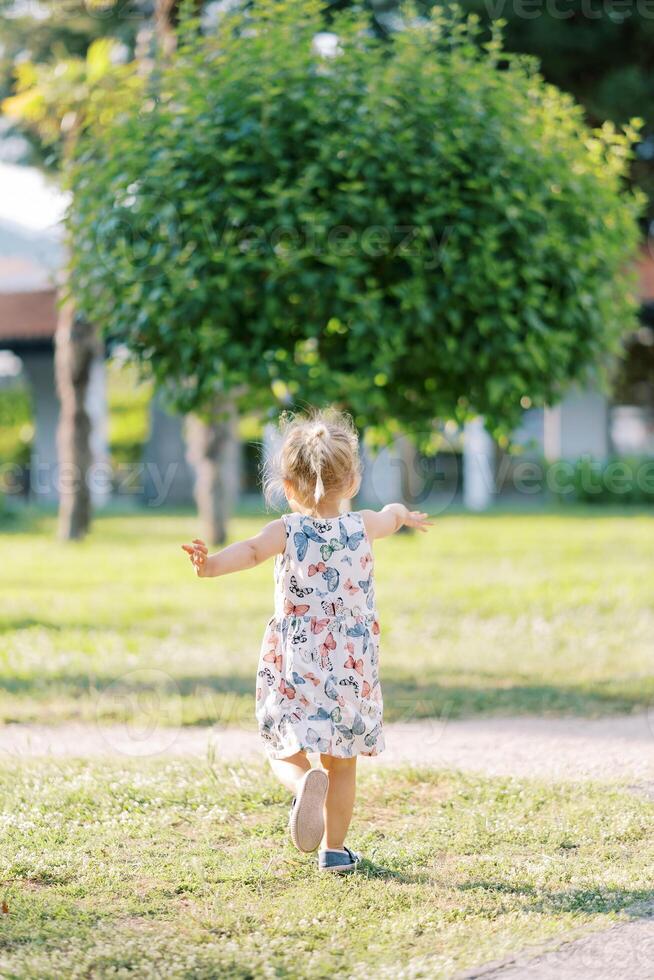 pequeño niña camina a través de un soleado césped hacia un verde árbol ondulación su brazos. espalda ver foto