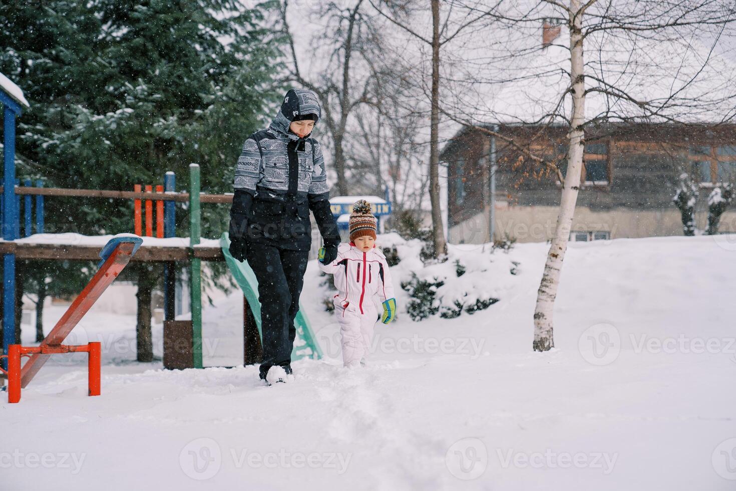 Mom and little daughter walk holding hands past a snowy slide under snowfall photo