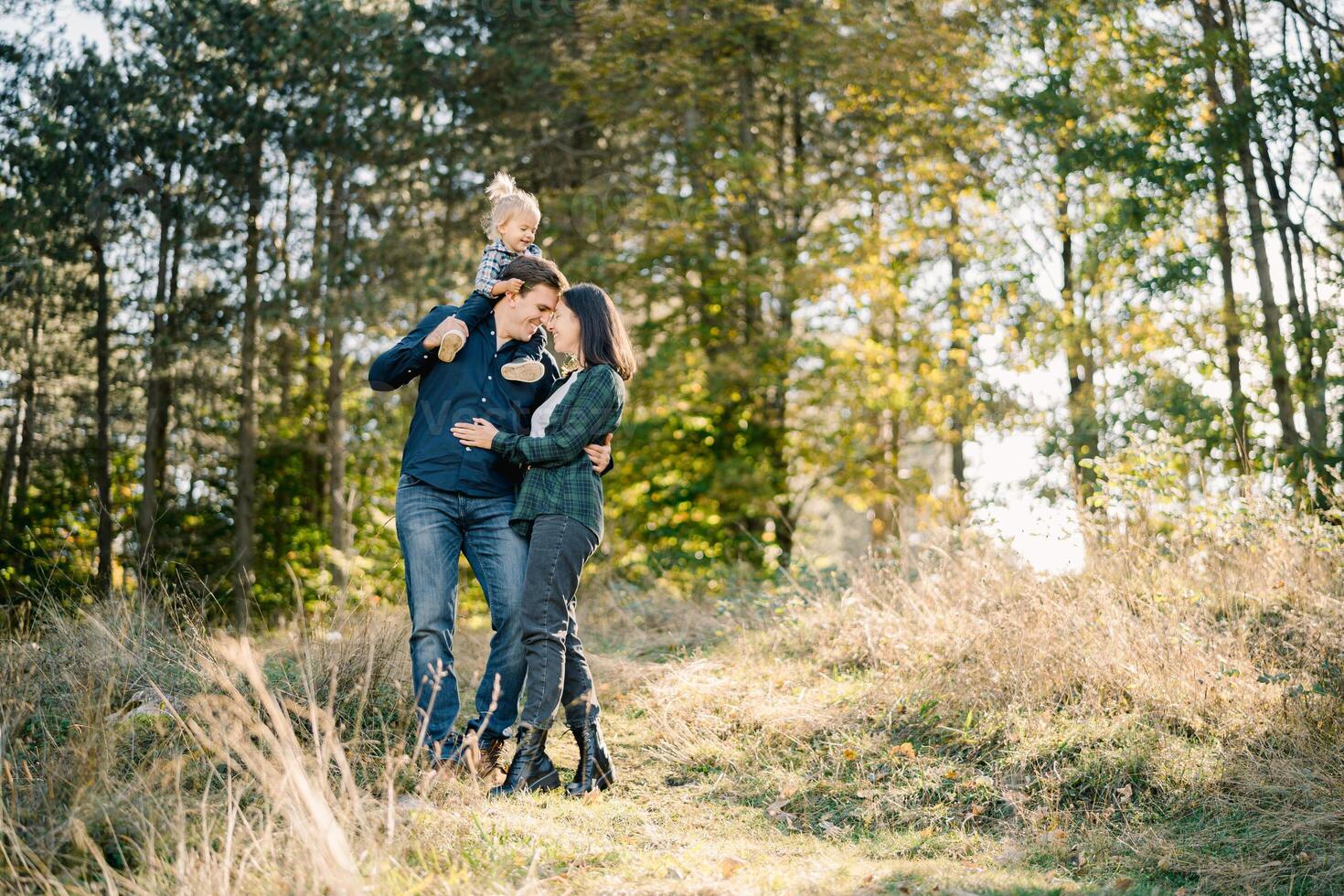 Mom hugs dad with a little girl on his shoulders touching his forehead with her forehead photo