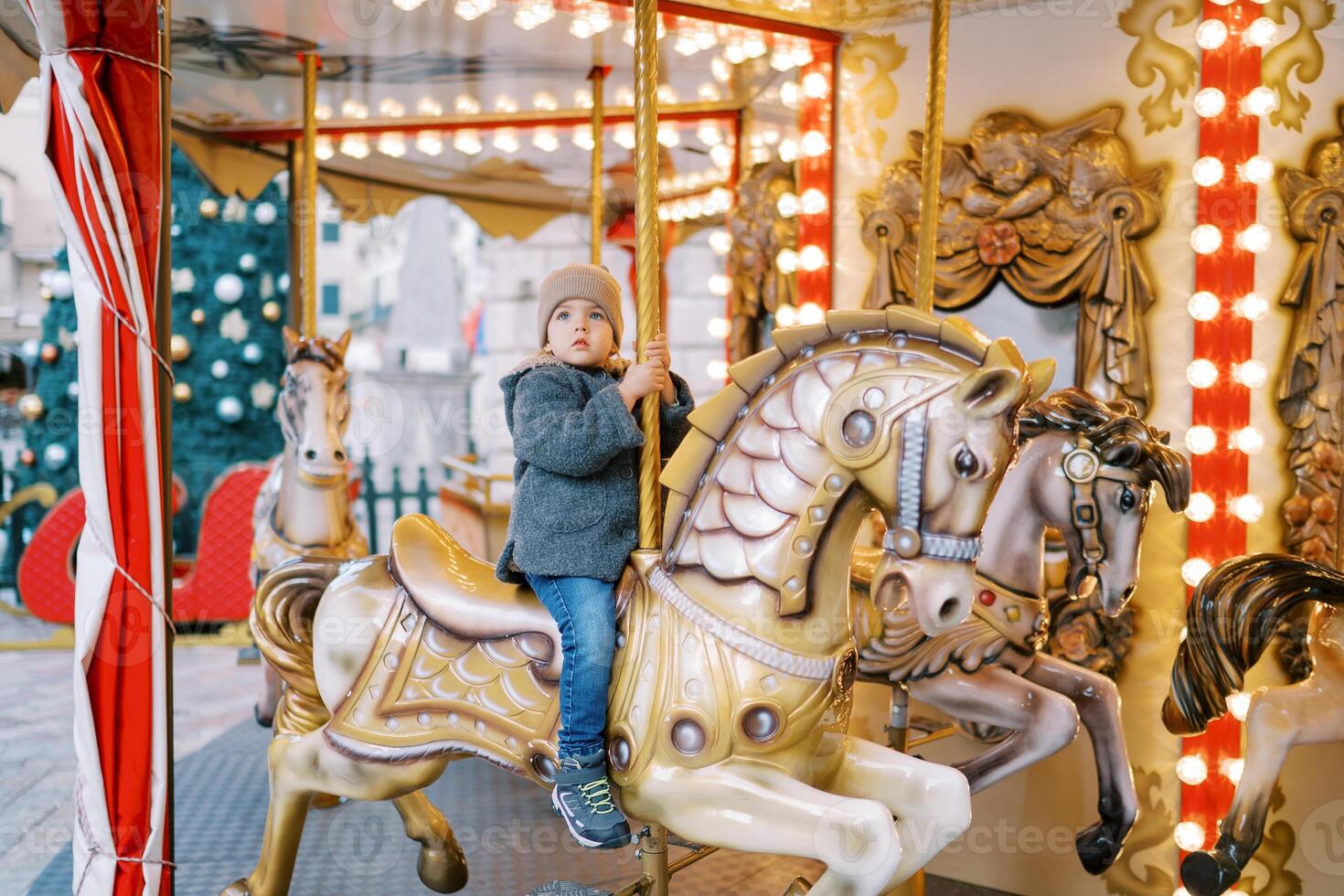 Little girl rides a toy horse on a carousel in the square near a decorated Christmas tree photo