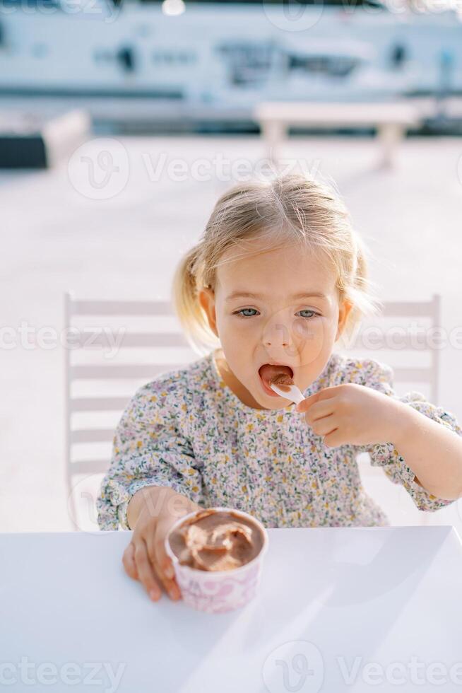 pequeño niña come chocolate hielo crema con un cuchara desde un taza mientras sentado a un mesa foto