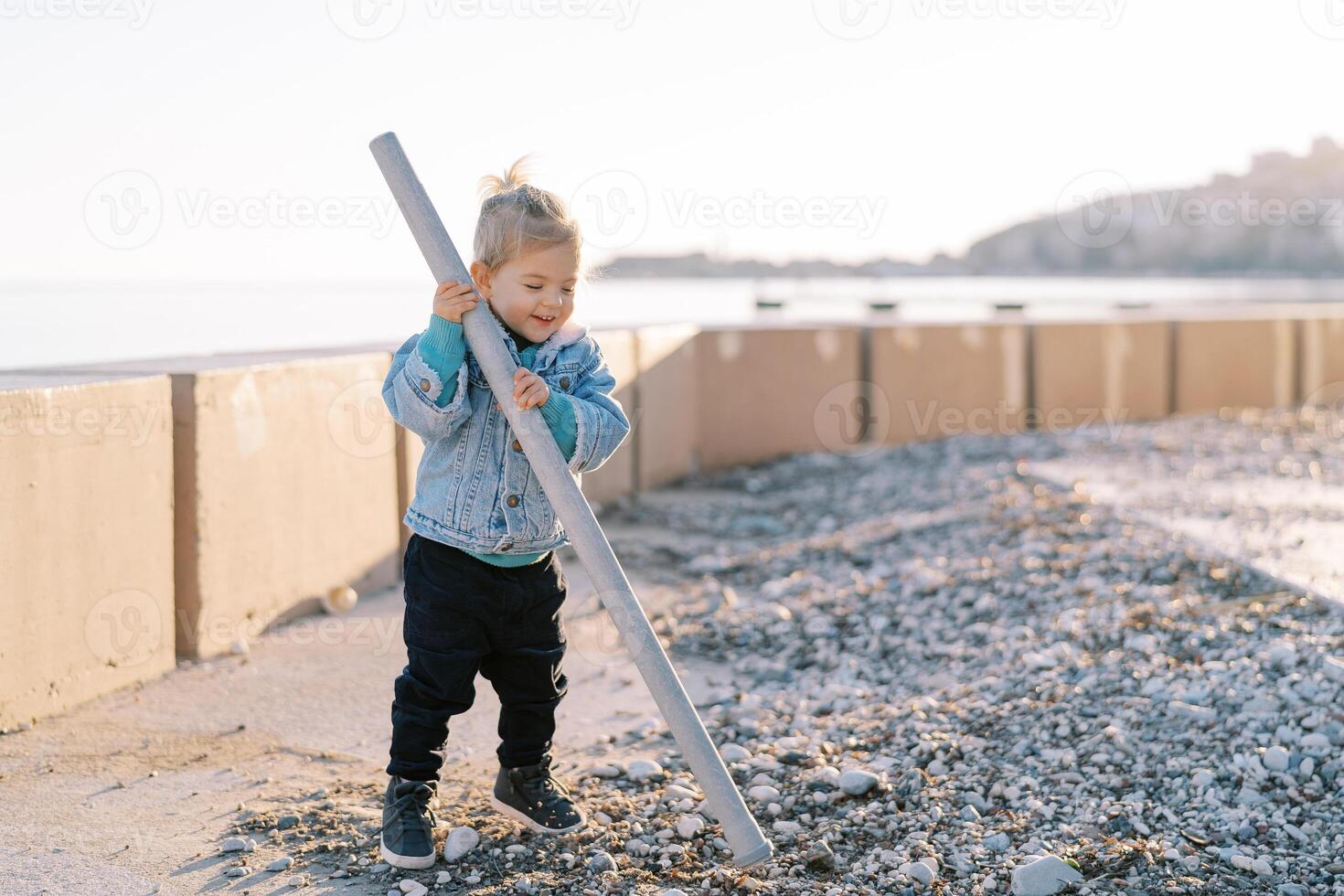 Little laughing girl picking pebbles with a plastic pipe while standing near the fence on the seashore photo