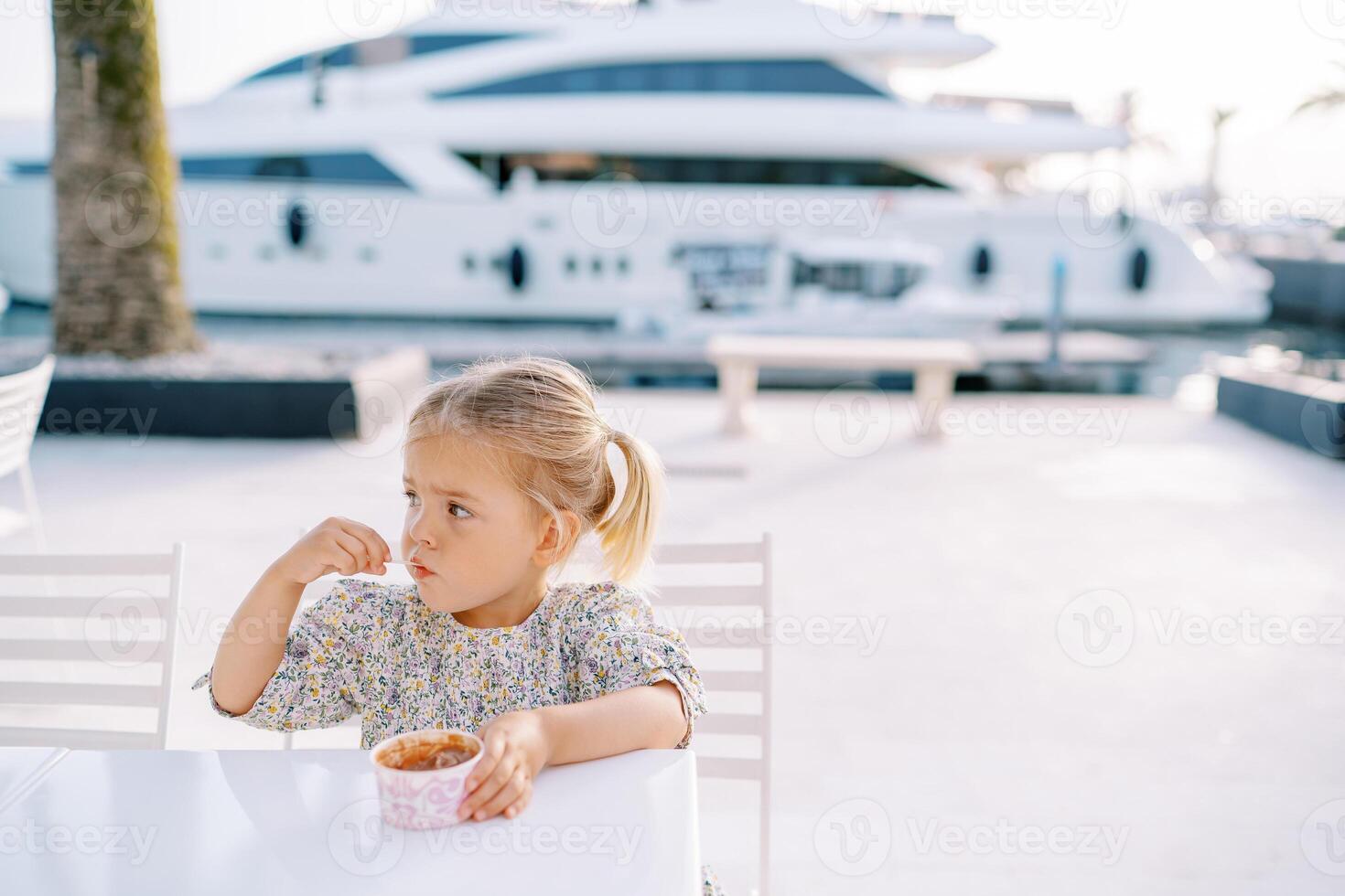 Little girl eats ice cream with a spoon, holding a glass with her hand on a table on the seashore photo