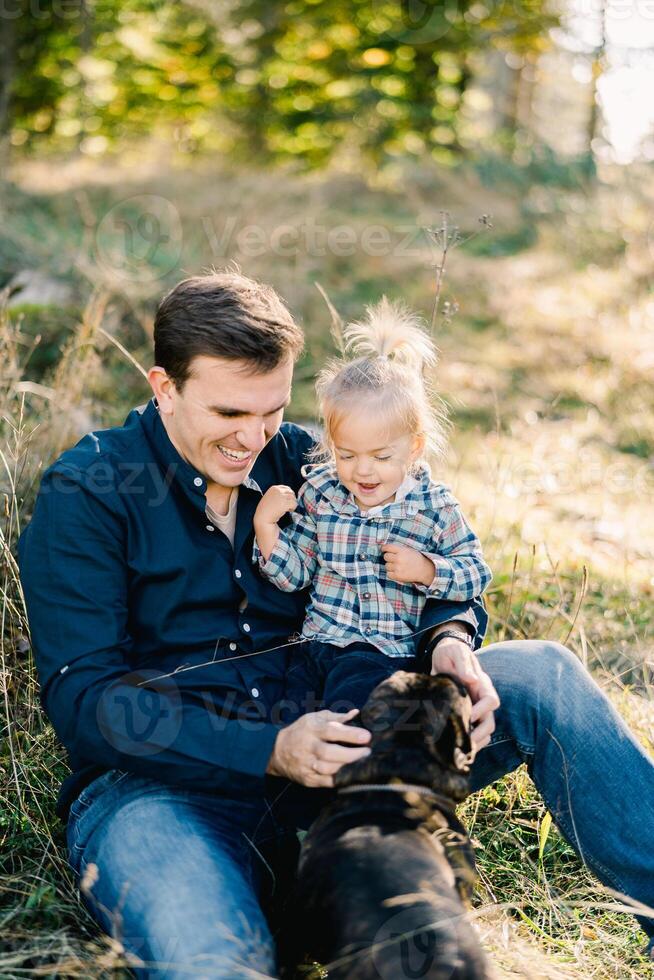 Dad sits on the lawn with his little daughter and strokes the dog photo