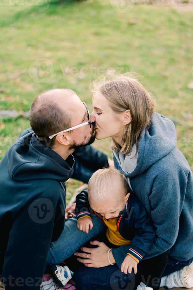 Dad kisses mom with a little boy on her lap while sitting on a green lawn photo