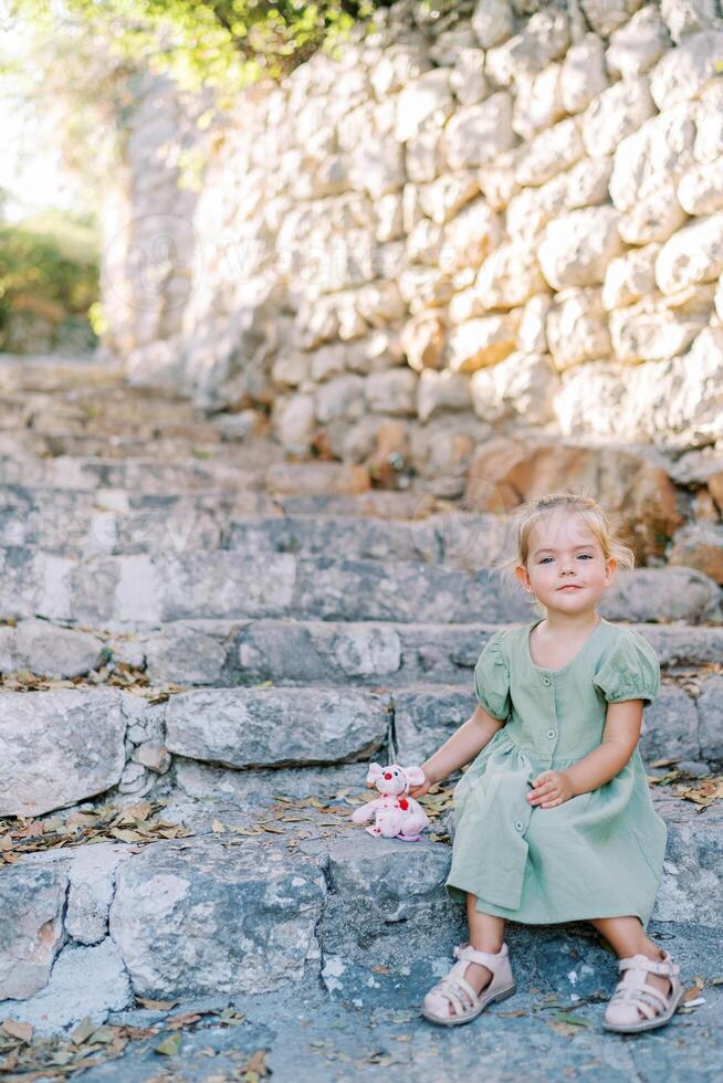 Little smiling girl with toy pink mouse sitting on old stone steps in the garden photo