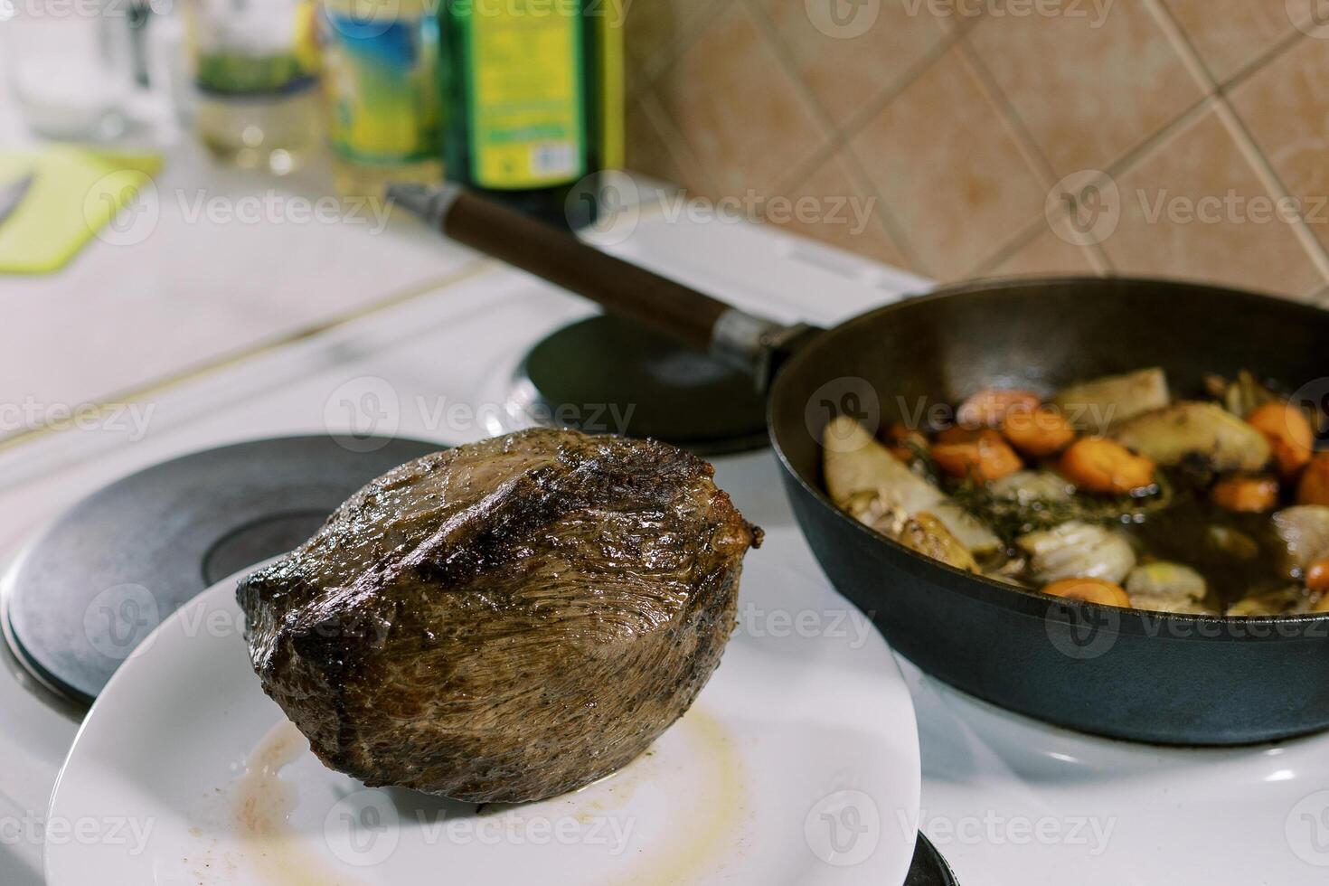 Baked tenderloin lies on a plate next to vegetables in a pan on the stove photo