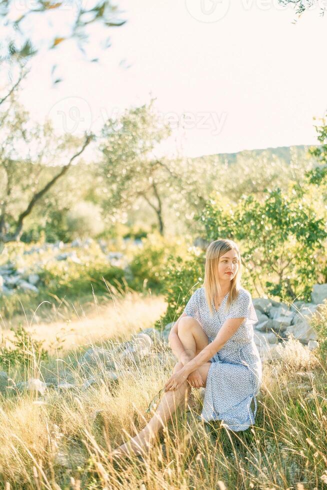 niña se sienta en el piedras en el parque, abrazando su elevado rodilla y mirando a el lado foto