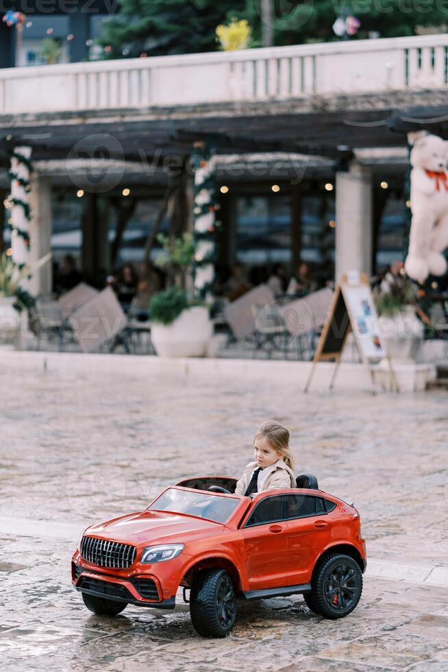 Little smiling girl drives a red toy car down the square photo